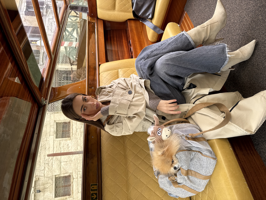 A female individual is seated inside a private water taxi or boat in Venice, Italy. She has long dark brown hair and is wearing a beige trench coat over a grey sweater, faded blue wide-leg jeans, and beige high-heeled boots. Her right hand is raised to her right temple, and she looks directly forward with a neutral expression. To her left, a small, fluffy, light brown Pomeranian-type dog is visible, resting inside a grey fabric pet carrier bag adorned with a small brown, crocheted animal charm. A black shoulder bag is on the seat to her right. The boat's interior features polished wooden ceilings and trim, mustard-yellow quilted leather benches, and large windows. Through the windows, traditional Venetian architecture is observable, including aged stone buildings with multiple windows, some with security bars, and sections of other structures with reddish elements, confirming an urban canal-side setting in Venice.