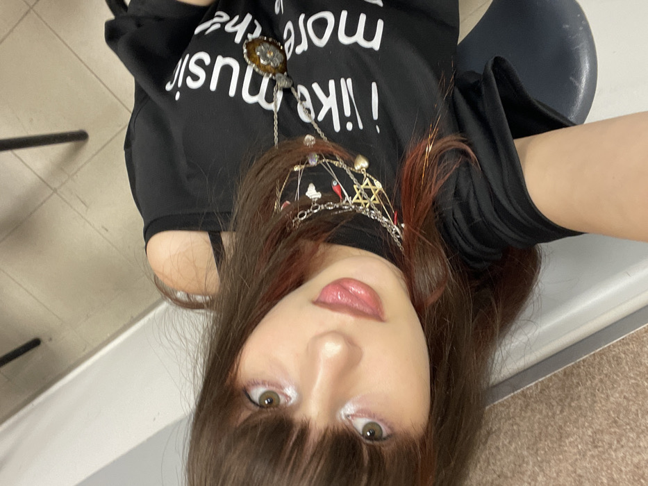 The image displays a close-up, angled portrait of a young female seated indoors, looking directly at the camera. She has long, dark brown hair with bangs and visible reddish-auburn highlights. Her eyes are greenish-brown, and she wears prominent white eyeliner on her lower lash line, along with a glossy, light pink lip product. She is wearing a black t-shirt with cut-out shoulders, revealing a black strap on her left shoulder. White text on the shirt reads "i like music" followed by partially visible "more th". Multiple layered necklaces adorn her neck, including a silver chain with a large, circular pendant featuring a brown, possibly amber-colored stone and a silver floral design, alongside other silver chains with various charms such as a gold-toned Star of David, small hearts, and red accents. The background, out of focus, shows a light-colored wall or partition to the left, a light beige or brown tiled floor, and the dark vertical leg of an object and a dark blue or grey seat to the right. The generic indoor setting offers no specific architectural or landmark identifiers to confirm a precise location within the city of Raanana, Israel.