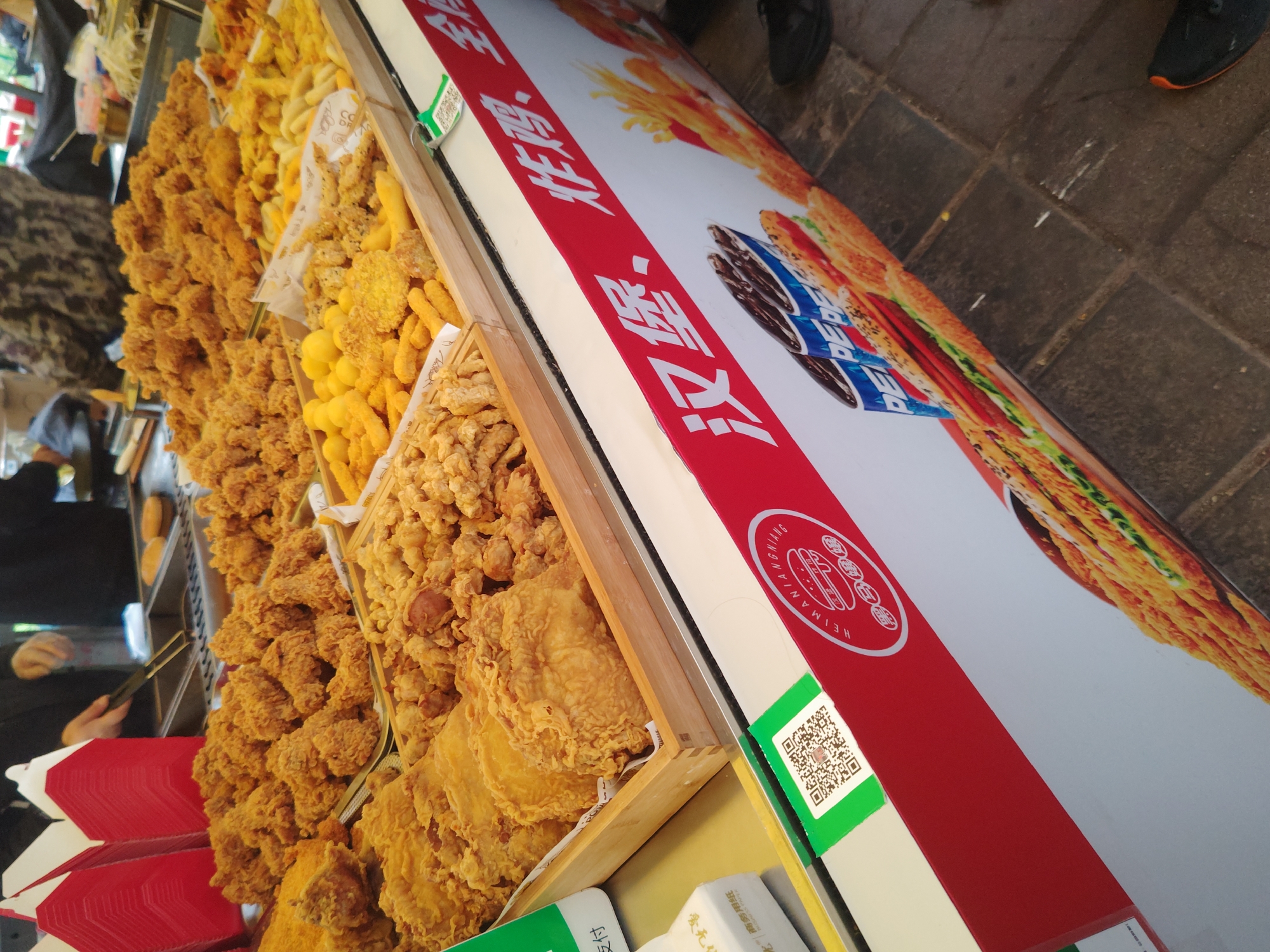 The image shows a food stall in Chongqing High-tech Zone, China, laden with various fried foods. The stall features a sign with red lettering and imagery of fried chicken and Pepsi products. The fried food items, arranged in wooden trays, include what appears to be chicken pieces, nuggets, and fried dough. A person is seen using tongs, likely serving food to customers. The stall is located outdoors, with a brick-paved walkway visible.