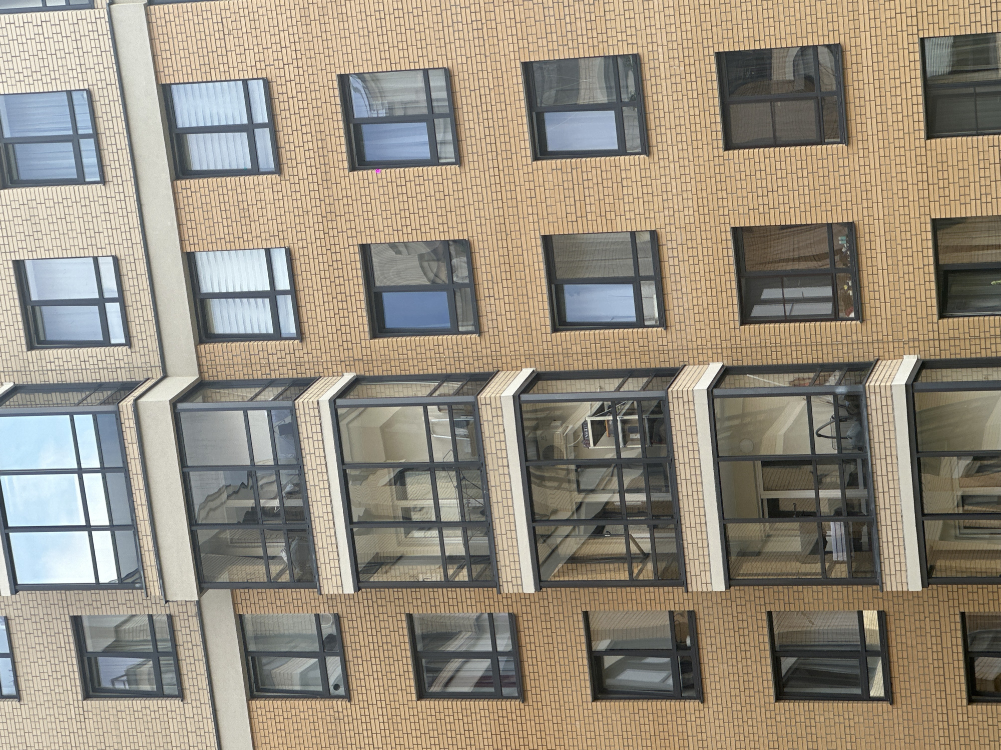 A close-up, eye-level, exterior, vertical shot shows the facade of a modern apartment building constructed from light brown bricks. The building features a prominent bay window structure on the left, comprised of multiple floors of glass and dark metal framing. Numerous other windows are visible across the facade, some with blinds or curtains. The lighting suggests it is daytime, with reflections of the sky and clouds visible in some of the glass panes. No people are visible in the image.