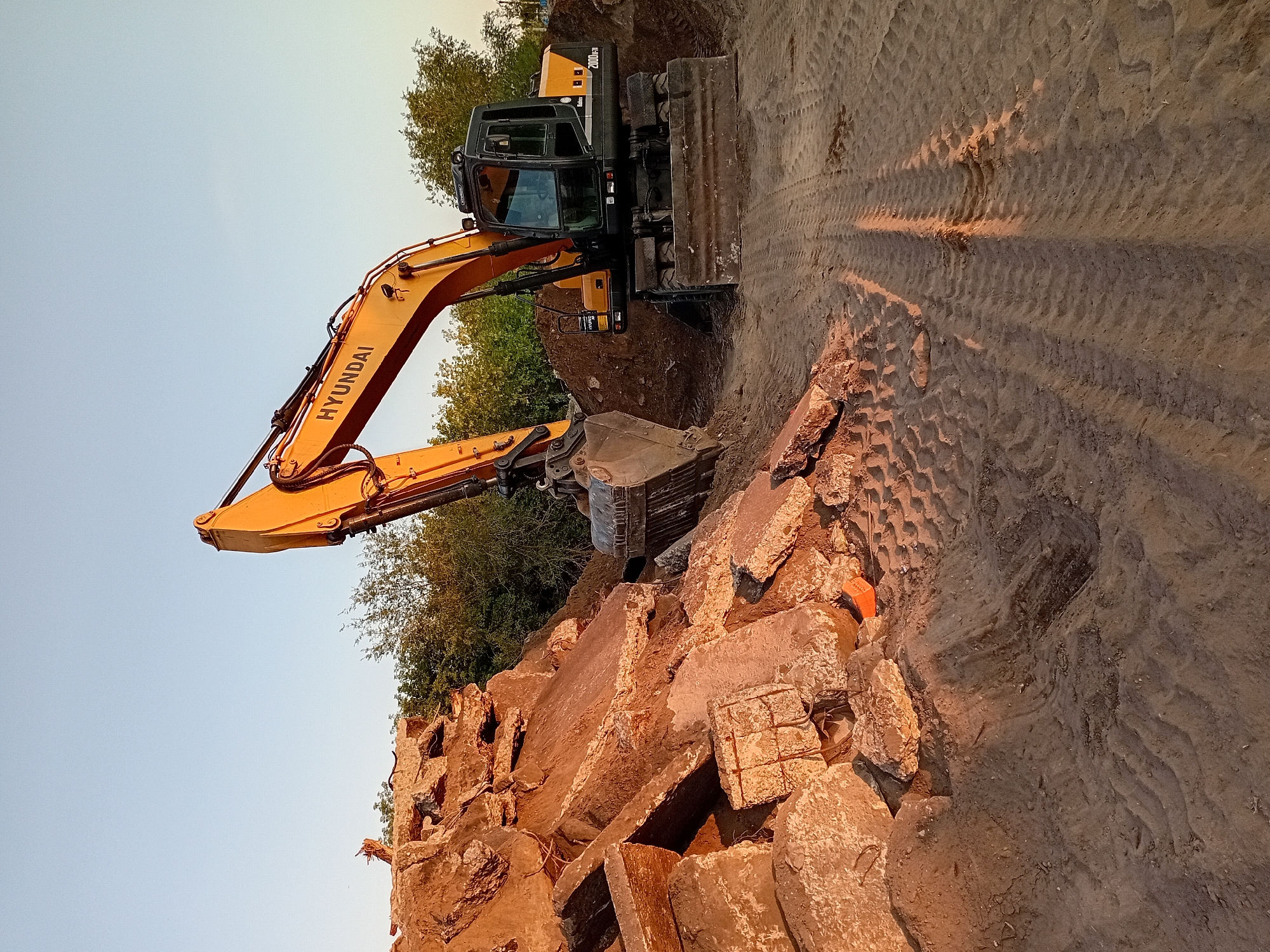 The image shows an orange Hyundai excavator in a construction site with piles of concrete debris and loose sand. The excavator is positioned to the right of the frame, with its bucket extended over a mound of earth. Tire tracks are visible in the sand, indicating recent activity. In the background, trees line the horizon under a clear blue sky. The lighting suggests it is late afternoon or early morning. The location of Semey, Kazakhstan, is not discernible from the visual information in the image. There are no people or discernible animals present in the scene.