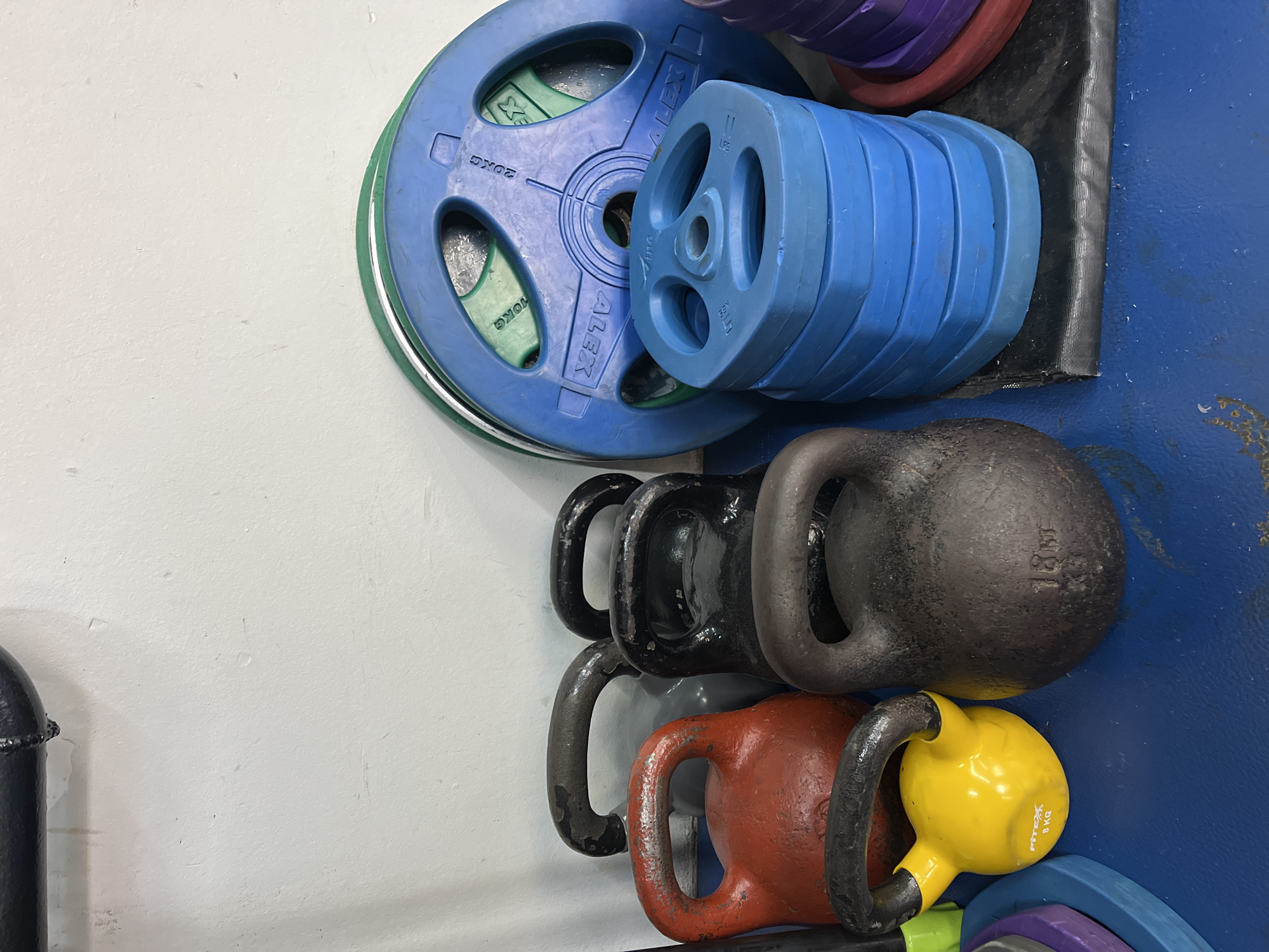 This media file captures a collection of gym equipment, specifically weight plates and kettlebells, arranged in a storage area. No people are visible.
The scene depicts a section of a gym or fitness facility, characterized by a plain white wall on the left and a blue surface (likely a floor or structural element of a weight rack) on the right. A black mat separates some of the equipment from the blue surface. The items appear to be stored vertically on racks or shelves against the wall.
In the upper section of the image, various weight plates are visible. A large blue plate, clearly marked "20KG" and "ALEX," dominates the left side, resting against smaller green plates (one of which shows "10KG"). To its right, a stack of smaller, lighter blue plates is arranged horizontally, with some displaying "LBS." Further right and slightly behind, purple and red weight plates are also discernible.
Below the weight plates, a series of kettlebells are stacked. These include a large black kettlebell, which has "18KG" embossed on its side. Below it, a red kettlebell is visible, and at the bottom of the stack, a bright yellow kettlebell is prominently displayed, bearing the text "Fitex 8 KG." Other kettlebells in black and grey are partially obscured.
The equipment shows signs of use, including scuffs, scratches, and some worn paint, indicating a functional and active gym environment. The arrangement suggests an organized storage system within a fitness setting in Krasnoyarsk, Russia.