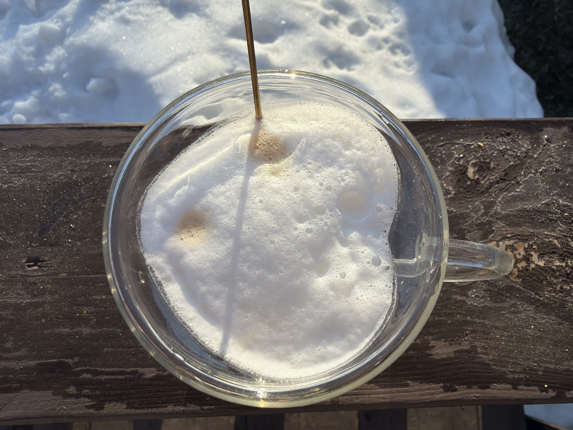 A top-down view shows a clear glass mug filled with a foamy, white beverage, likely a cappuccino or latte. A thin, brown-colored stick or straw is inserted into the center of the foam. The mug sits on a weathered, dark brown wooden surface, possibly a deck or railing. In the background, there is a patch of snow. It is highly unlikely, based solely on the image, to ascertain the location as the city of Chaikovsky, Russia.