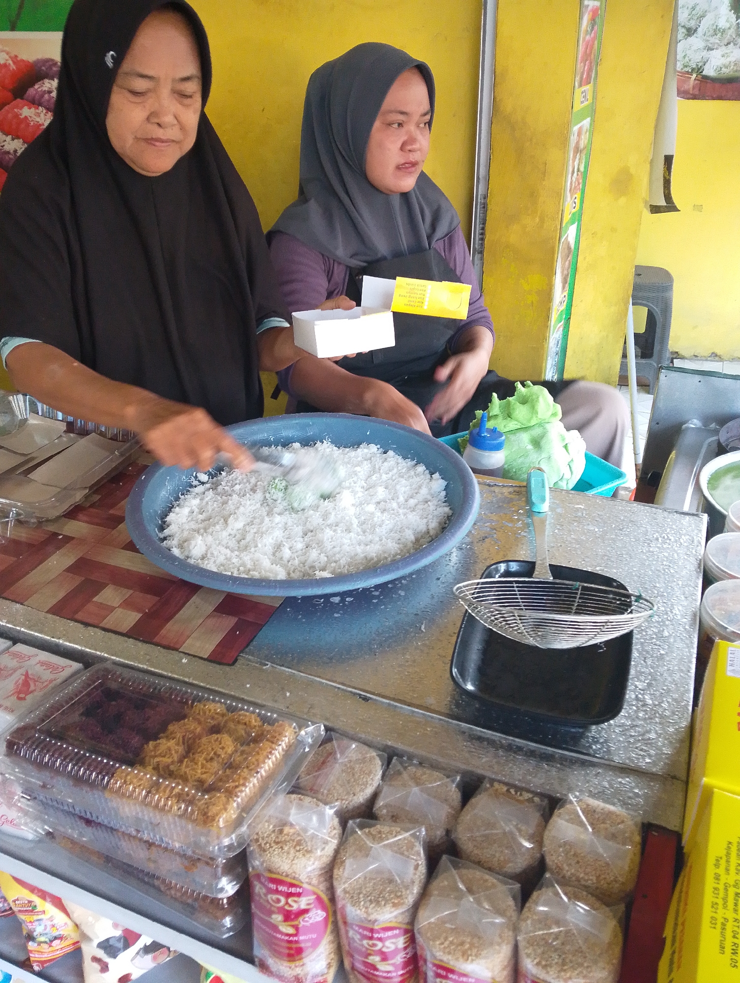In Legok, Indonesia, two women are preparing food at a street stall. The woman on the left, wearing a black hijab, is mixing shredded coconut in a blue plastic basin with a scoop. To her right, a woman wearing a grey hijab holds a small white box. Behind them, a yellow wall displays various food items. In the foreground, a metal strainer sits on a black tray. Containers of snacks are visible on the counter, including what appear to be sweet snacks and bags of sesame seeds. The scene suggests a busy food preparation environment at a local market or roadside stall.