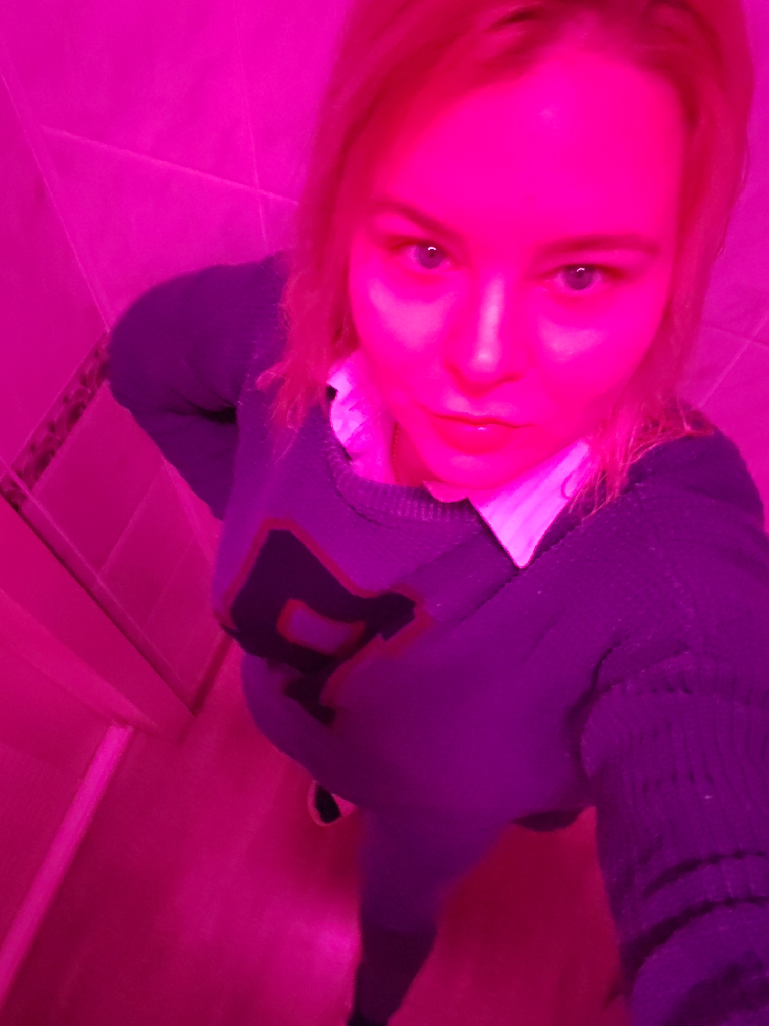 A young woman with blond hair and blue eyes is taking a selfie in a room with pink lighting. She is wearing a gray sweater with a red and black "P" on it and a white collared shirt underneath. She appears to be standing in a bathroom, based on the tiled wall visible in the background. The location in Izhevsk, Russia, is not discernible from the image.