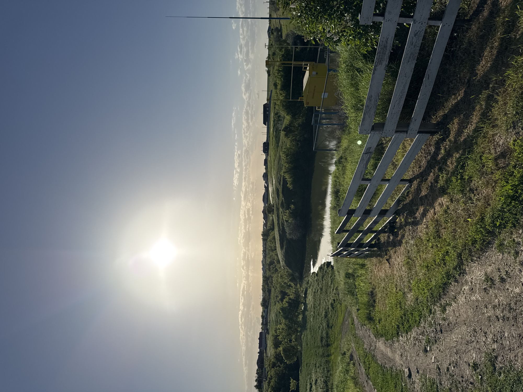 The image depicts a rural landscape in Усть-Курдюм, Russia, under a bright, hazy sky with the sun prominently visible. A body of water, possibly a river or canal, runs through the scene, reflecting the sunlight. A white, wooden fence is situated on the right side of the frame, casting shadows on the grassy bank. A dirt path follows the contour of the land alongside the water. Across the water, dense vegetation and some visible infrastructure, including a yellow structure and utility poles, are present. In the distance, rooftops of houses are visible amidst the trees.