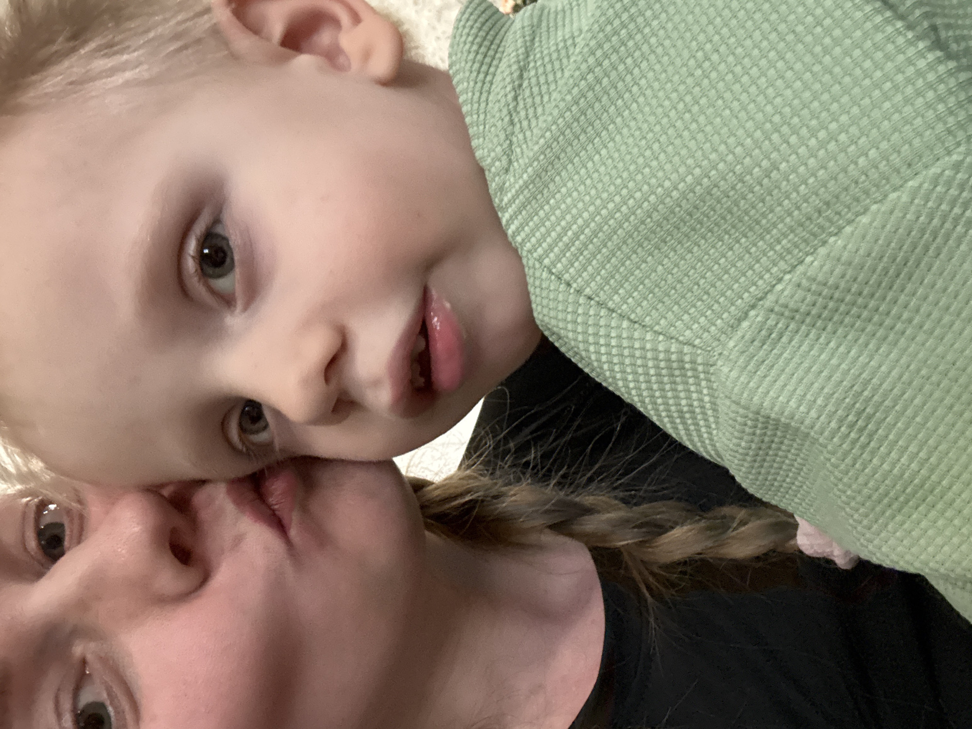 A close-up shot features a child and an adult, likely a parent, in an intimate pose. The child, with light blonde hair, fair skin, and grey eyes, is positioned above the adult. The child is wearing a light green textured jacket. The adult has blonde hair braided in a single plait, fair skin, and grey eyes. The adult wears a black shirt. Their faces are pressed together, suggesting a kiss. There is nothing in the image to suggest that the image was taken in the city of Chaikovsky, Russia.