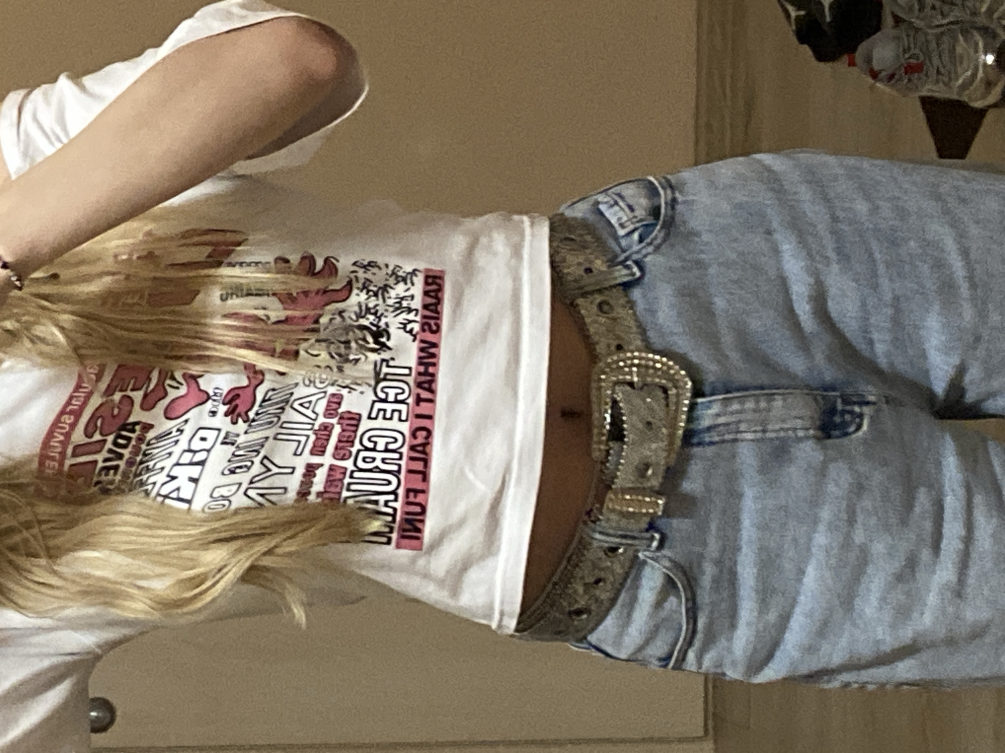A waist-up shot depicts a person wearing a white t-shirt with pink and black text. The text is largely illegible due to being partially obscured by blonde hair and being printed in reverse. However, a portion of the text reads "ICE CREAM FUN". The person is also wearing light-wash blue jeans with a bejeweled belt. The background is a plain, neutral-colored wall. The location appears to be indoors.