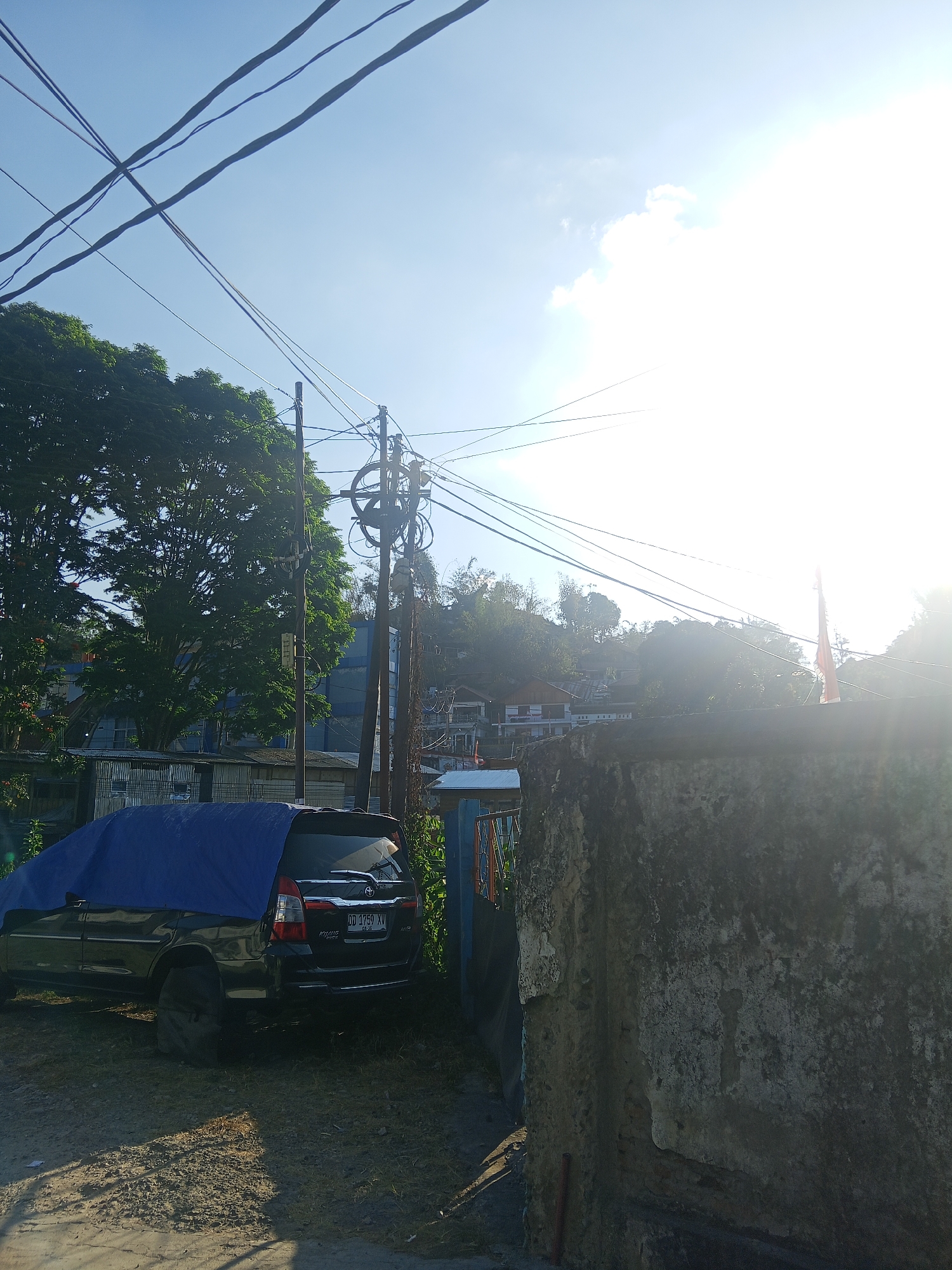 In Makale, Indonesia, a black SUV is parked on a dirt lot, partially covered by a blue tarp. The vehicle's license plate reads "DD 1759 XV". The area appears to be a residential neighborhood with houses visible on a hill in the background. Overhead, a tangle of power lines crisscrosses the sky, silhouetted against a bright, sunny sky with some clouds. A large tree with dense foliage stands to the left of the vehicle, and a weathered concrete wall occupies the right foreground. The scene suggests a daytime setting with strong sunlight casting shadows on the ground.