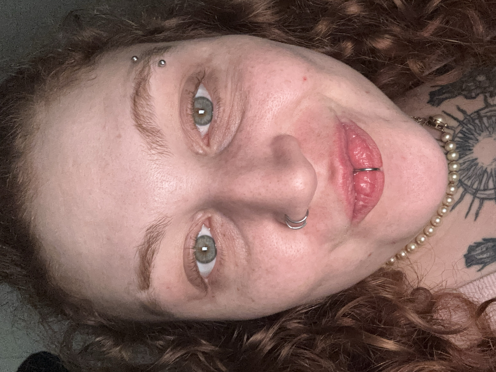 A close-up, slightly angled, portrait shot depicts a person with fair skin, red curly hair, and distinct facial piercings. The person has an eyebrow piercing, a nose piercing, and a lip piercing. Their eyes are a light, muted green color. The individual's skin appears to have freckles and some redness. They wear a pearl necklace. A tattoo is visible on the upper chest area. The person is not in Buenos Aires, Argentina. The background is blurry and nondescript.