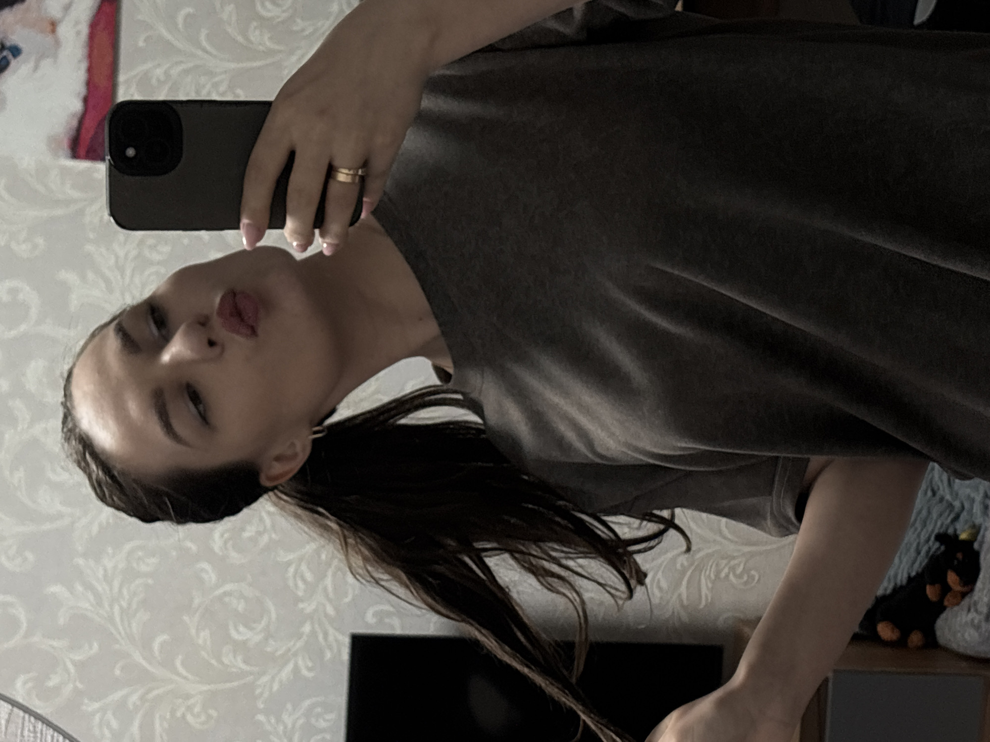 A person is taking a selfie. The person has dark hair in a low ponytail and is wearing a gray t-shirt. They are holding a smartphone with a black case, and a gold ring is visible on their ring finger. The background includes a wall with patterned wallpaper, a television, and a stuffed animal on a shelf. There is a partial view of a fan on the left. The location is not specified.