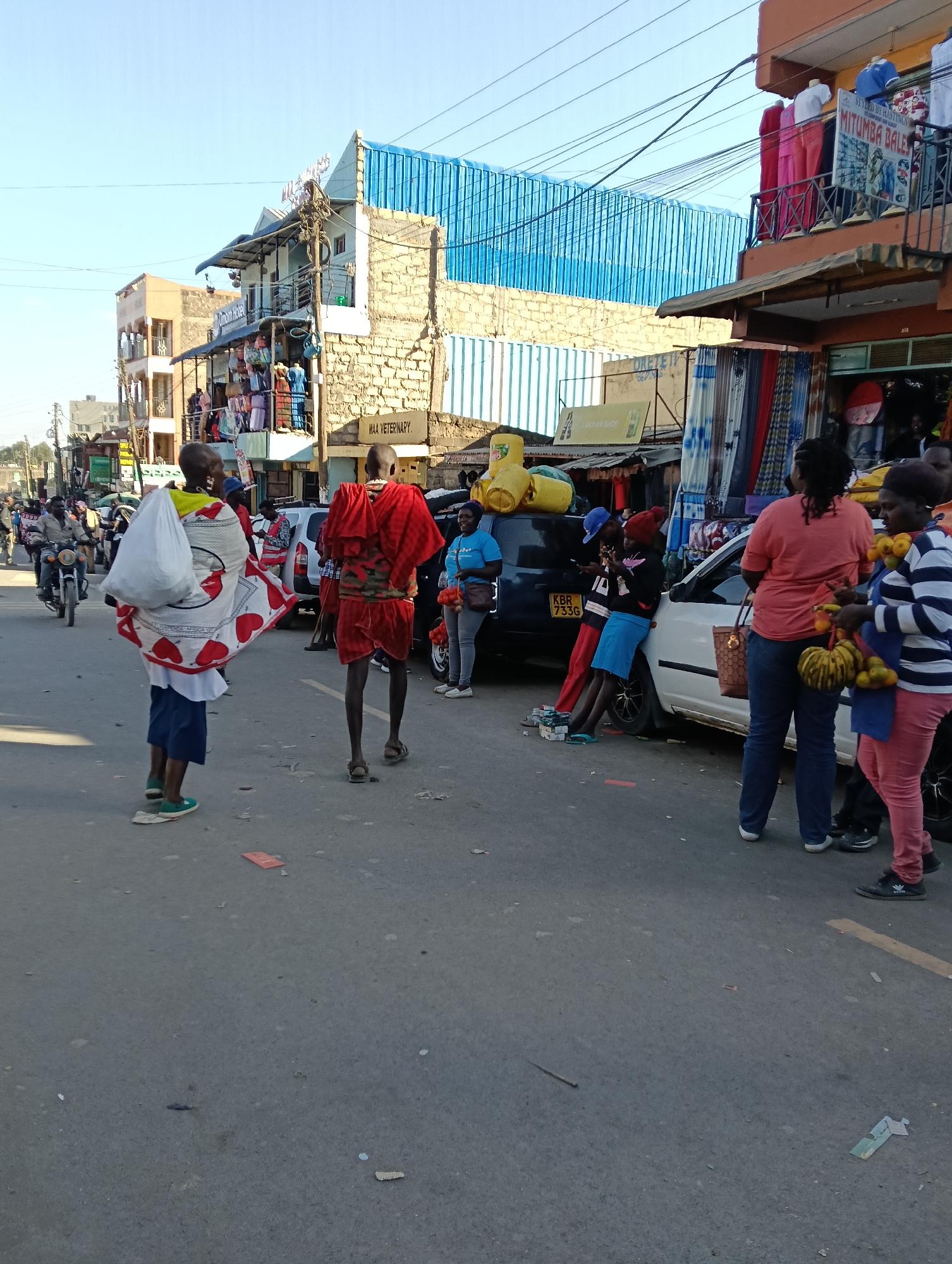 The image depicts a street scene in Keekonyokie Ward, Kenya, characterized by a bustling marketplace. In the foreground, two individuals are walking away from the camera. The person on the left is carrying a large white bundle wrapped in a red and white patterned cloth. The person on the right is wearing a traditional red garment and a patterned fabric around their torso. Several people are gathered around cars parked on the street. One car, a white sedan with license plate KBR 733G, has individuals leaning against it. Another dark-colored car is parked behind it, with yellow containers stacked on top. Shops line the street, with displays of clothing and goods visible. A motorcycle is seen in the background, heading down the street. The environment appears to be an outdoor marketplace with a clear blue sky overhead and buildings with multiple stories forming the backdrop. The ground is asphalt, with some debris scattered on it.