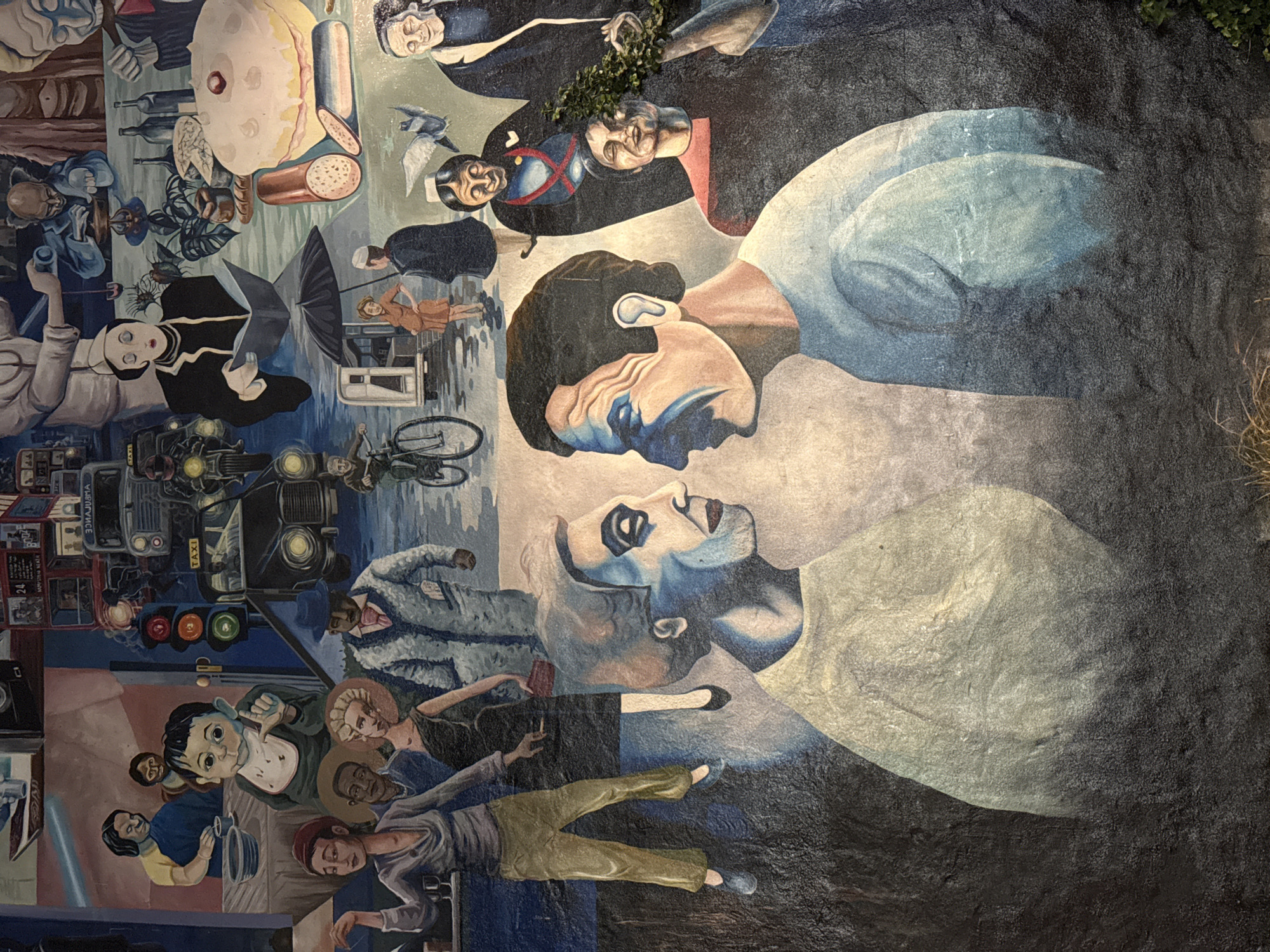 The image is of a section of the "Fitzrovia Mural" in London, United Kingdom. The mural section depicts a complex layering of scenes and characters, creating a surreal and dreamlike atmosphere.
The central focus is on two large, stylized faces looking at each other, rendered in a slightly Cubist manner with prominent features. Below them is a scene with figures in various poses, including a person lying down in olive-green pants, and other standing. To the left, there's a collection of objects and figures, including a red double-decker bus, a taxi, people under umbrellas, a cyclist, and a table with food and bottles. The composition is densely packed, with figures and objects overlapping. The mural is painted in a muted palette with grays, blues, browns, and whites dominating. The mural is partially obscured by vegetation.