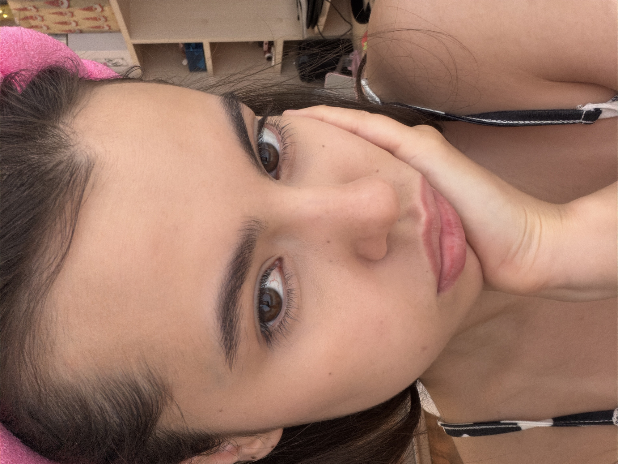 A close-up, rotated view of a young woman's face. She has dark brown hair, light skin, and dark brown eyes. She is wearing a black and white patterned bikini top and has a pink head wrap or towel on her head. Her left hand is resting against her cheek. The background appears to be a shelf or storage unit. There is no explicit indication of this image being taken in Chaikovsky, Russia.