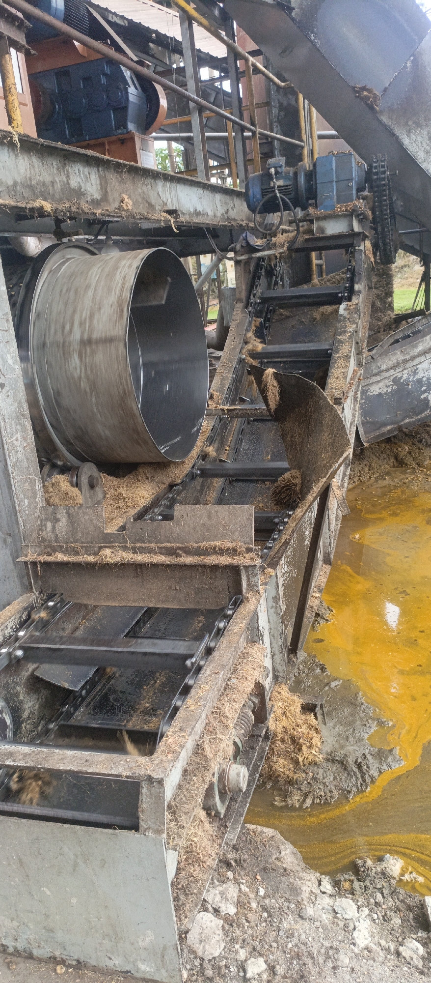 The image depicts an industrial scene, likely a processing facility, with heavy machinery and fibrous material. In the foreground, a large metal drum is visible, appearing to be part of a conveyor or processing system. Surrounding the machinery is a significant amount of light brown, fibrous material, which appears to be waste or raw material. There is also a pool of yellowish-orange liquid in the lower right portion of the image, with mud and debris around it. The overall impression is one of an active, possibly messy, industrial operation.
Without additional context or specific landmarks, it is not possible to pinpoint the exact location within Iemirah, Indonesia. However, the scene suggests a facility involved in the processing of agricultural products, such as palm oil or similar fibrous crops, which are common in Indonesia. The presence of industrial equipment, waste material, and a processing area aligns with the types of operations found in such regions.