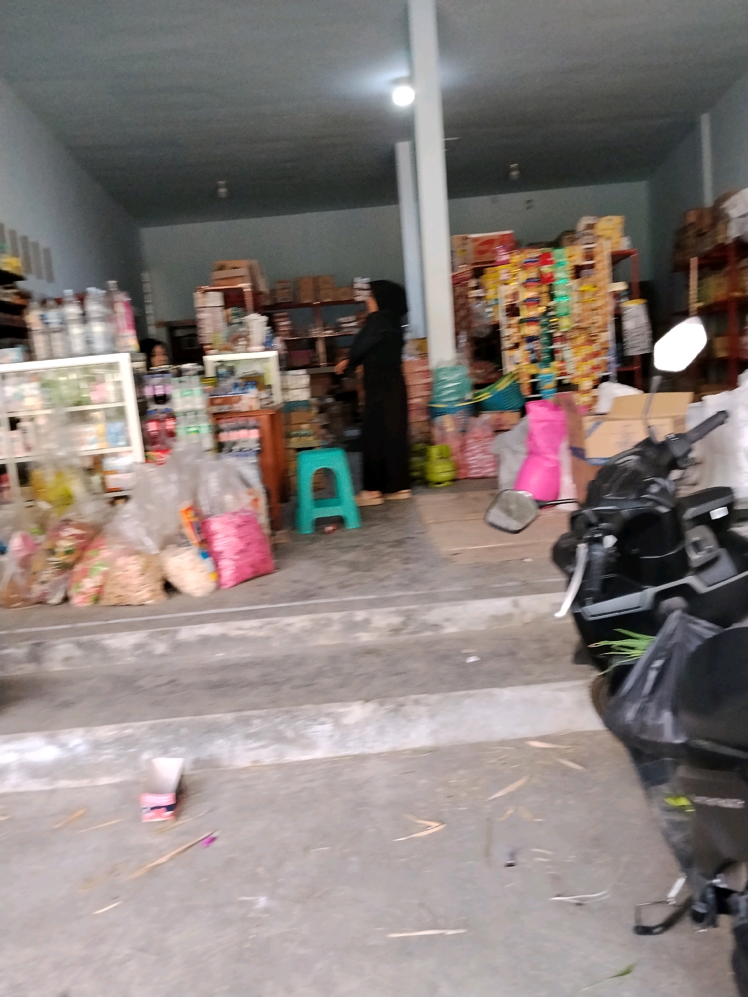 The image depicts the interior of a small convenience store or warung, located on a street in Panggungan, Indonesia. The store is stocked with various goods, including bottled drinks, packaged snacks, and household items displayed on shelves and in baskets. A person, possibly the store owner or an employee, is seen standing behind a counter, wearing a black hijab and black clothing. The floor is concrete, and there are several plastic bags filled with merchandise placed near the entrance. Outside the store, two motorcycles are parked, suggesting this is a typical street-level establishment. The overall scene conveys a sense of everyday commerce in a local Indonesian community.