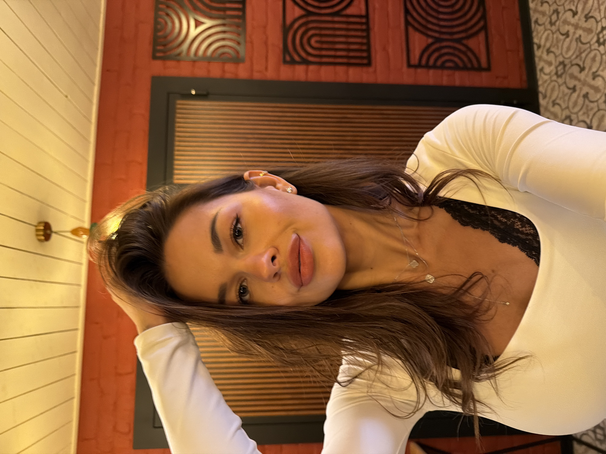 A young woman with long, dark hair is captured in a selfie taken indoors. She is wearing a white long-sleeved top with black lace visible at the neckline, and a delicate silver necklace. Her left arm is raised, with her hand touching her hair. The background features a reddish-orange brick wall with decorative metal panels, a wooden door, and a patterned floor. The lighting suggests an interior setting, possibly a restaurant or home. The specific location in Saint Petersburg, Russia cannot be determined from the image alone.