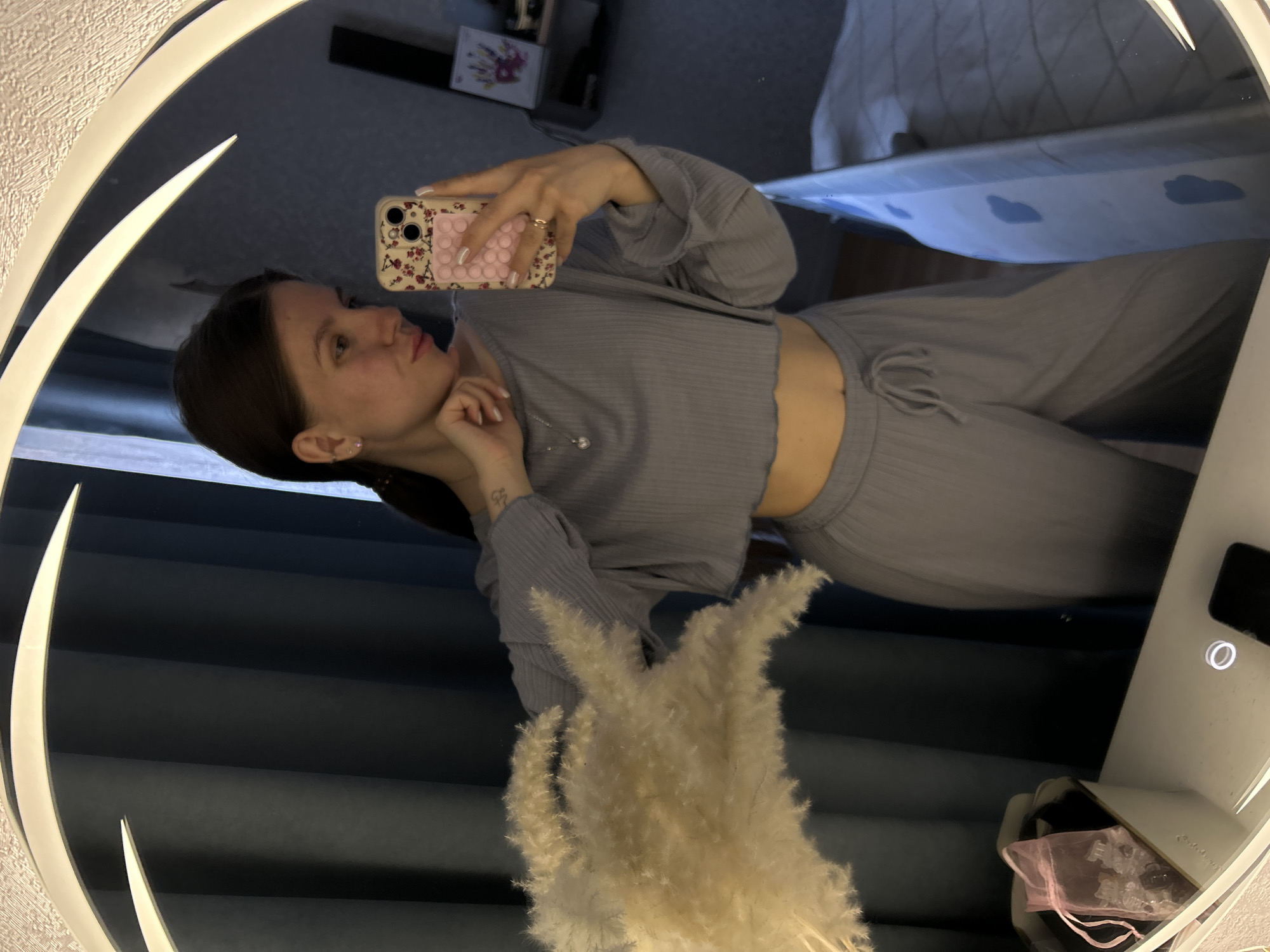 A person with dark hair is reflected in a round, illuminated mirror while taking a selfie. The individual is wearing a gray, two-piece ribbed outfit consisting of a cropped top and pants. They are holding a smartphone with a patterned case, pink and floral design and a pink phone grip or stand is visible on the back. The setting is a room with gray curtains and a glimpse of a bed with blue patterned bedsheets. A vase with decorative, white fluffy plumes is positioned in front of the mirror. The city of Klin, Russia is not visible in the image.