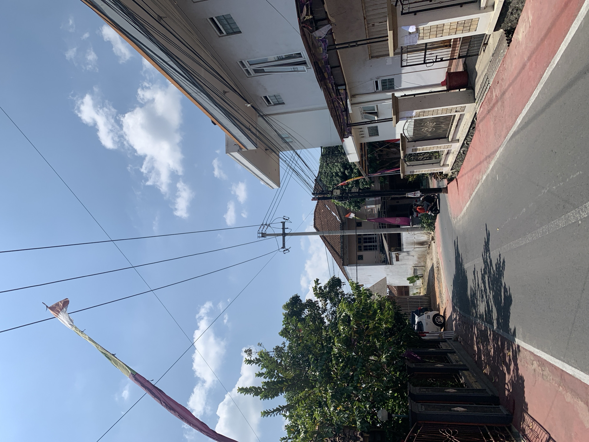 A medium shot captures a sun-drenched street scene in Ciamis, Indonesia, on a clear day with scattered white clouds. The road, paved with asphalt, is bordered by a vibrant red pedestrian walkway on the left. A tall, slender white car is parked along the side of the road, casting a shadow. To the left of the road, a dense green tree provides shade, its branches reaching towards the sky. Behind the tree, a dark metal fence with decorative elements delineates the property line. A power pole with multiple wires stretches across the frame, connecting to a white building with several windows. The building, an office or residential structure, has a modern design with clean lines and an orange-trimmed roof. On the right side of the street, a similar white building with an awning extends over the walkway. This building also features a metal fence with ornate designs. In the distance, more buildings with terracotta roofs are visible, suggesting a residential neighborhood. A colorful flag, likely a banner or a traditional Indonesian pennant, flies high on a pole, its fabric rippling in the breeze. The overall impression is one of a typical urban street in a tropical climate, with clear skies, lush vegetation, and functional architecture.