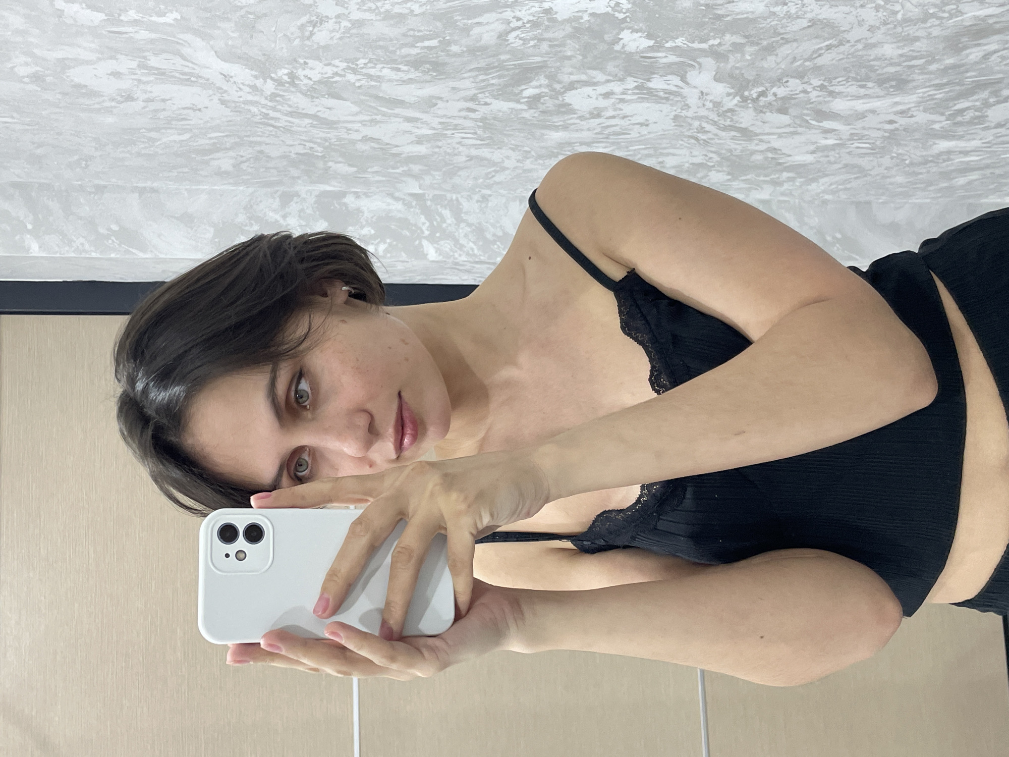 A woman is taking a selfie with a white smartphone. She has short, dark hair and is wearing a black camisole with lace trim. The background consists of a textured white ceiling and a beige wall. The location, Izhevsk, Russia, cannot be verified from this image alone.
