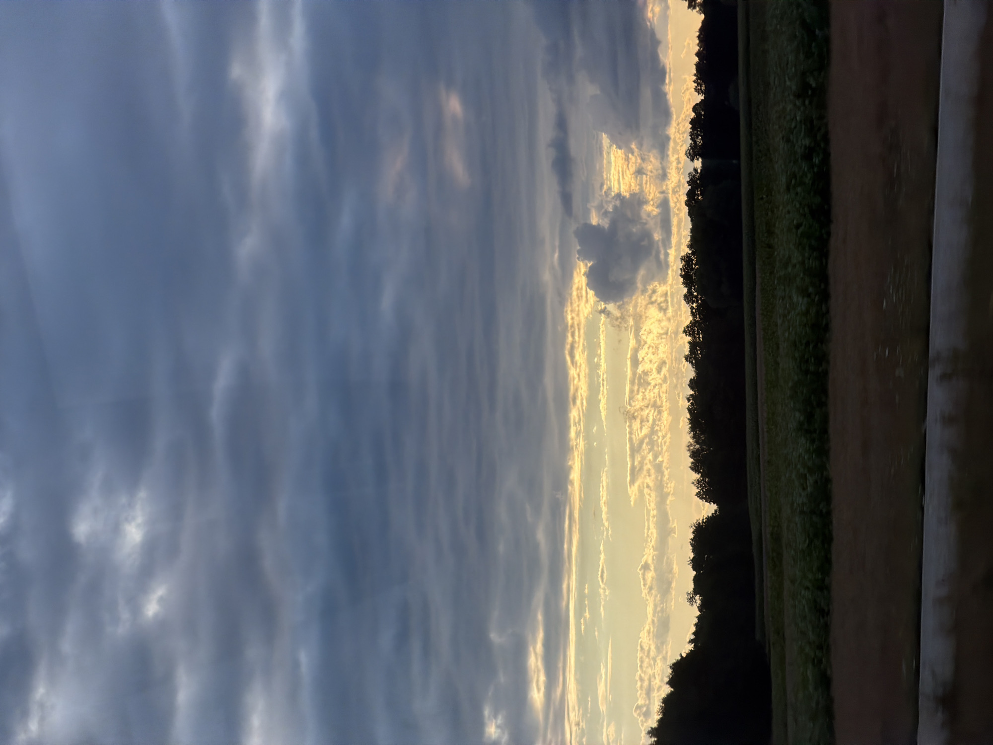 This is a full shot of a cloudy sky over a rural landscape in Kopaniny, Poland. The sky is filled with layers of dark gray and blue clouds, with some lighter patches of yellow and white peeking through, suggesting either sunrise or sunset. Below the sky, a dark line of trees runs horizontally across the image, forming the horizon. In front of the trees, a field of green crops extends forward, and further down, a strip of brown land is visible, possibly a dirt road or another cultivated field. The overall impression is one of a serene, natural setting under a dramatic sky.
