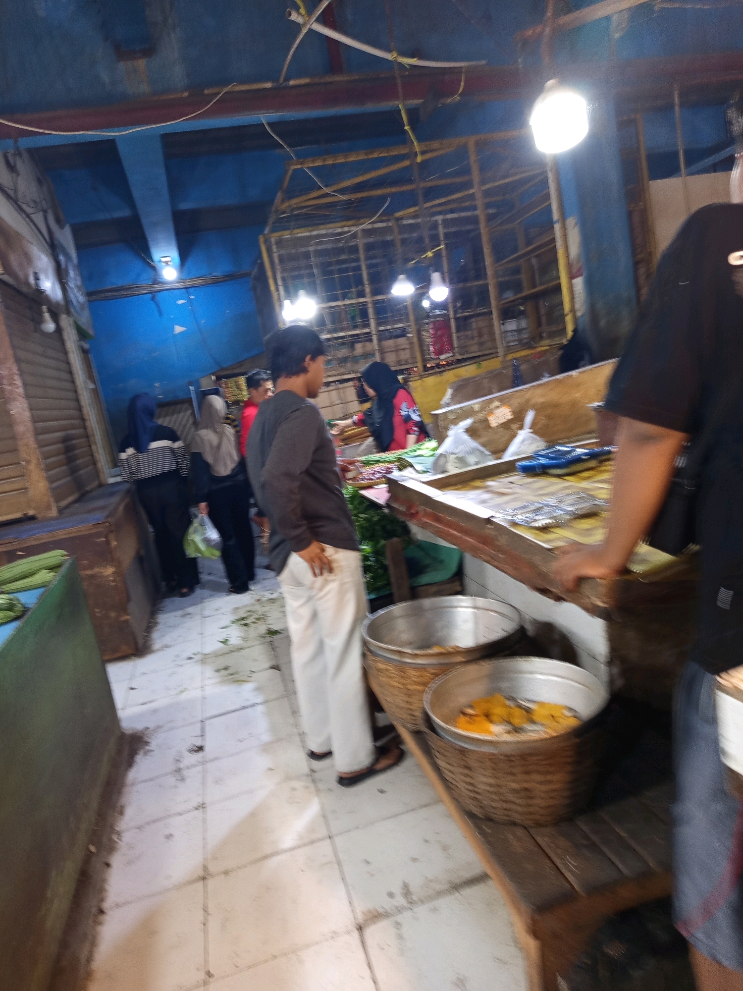 The image depicts an indoor market scene, likely in Karawang, Indonesia, illuminated by overhead lights. In the foreground, a man wearing a dark long-sleeved shirt and light-colored pants stands with his hand on his hip, facing away from the camera. To his right, a stall is visible with a wooden counter holding various items, including what appear to be packaged goods and possibly seafood. Two large woven baskets, one containing yellow food items, are placed on a wooden bench in front of the stall. In the background, several individuals, including women wearing headscarves, are browsing or working at other stalls. The market environment appears somewhat rustic, with exposed ceiling structures and painted blue walls. The floor is tiled and shows some scattered debris.