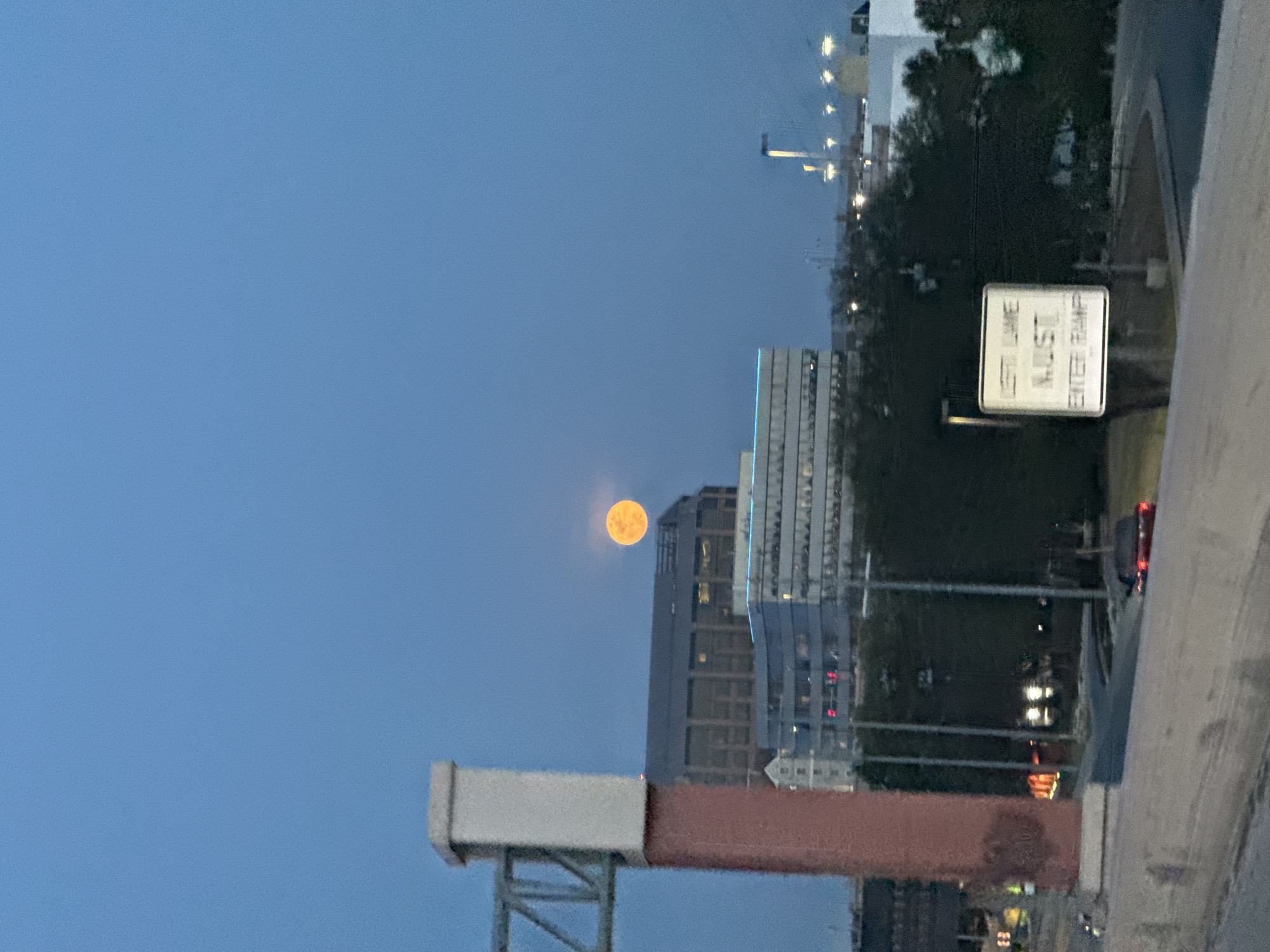 The image shows a view of San Antonio, Texas, at dusk, with the moon visible in the sky above the city buildings. A street sign indicating "Left Lane Must Enter Ramp" is on the right. The image was likely taken from an elevated position, such as an overpass or tall building. The architecture of the buildings suggests a modern urban setting. There are no visible people, but vehicles are present on the road. The sky is a light blue.