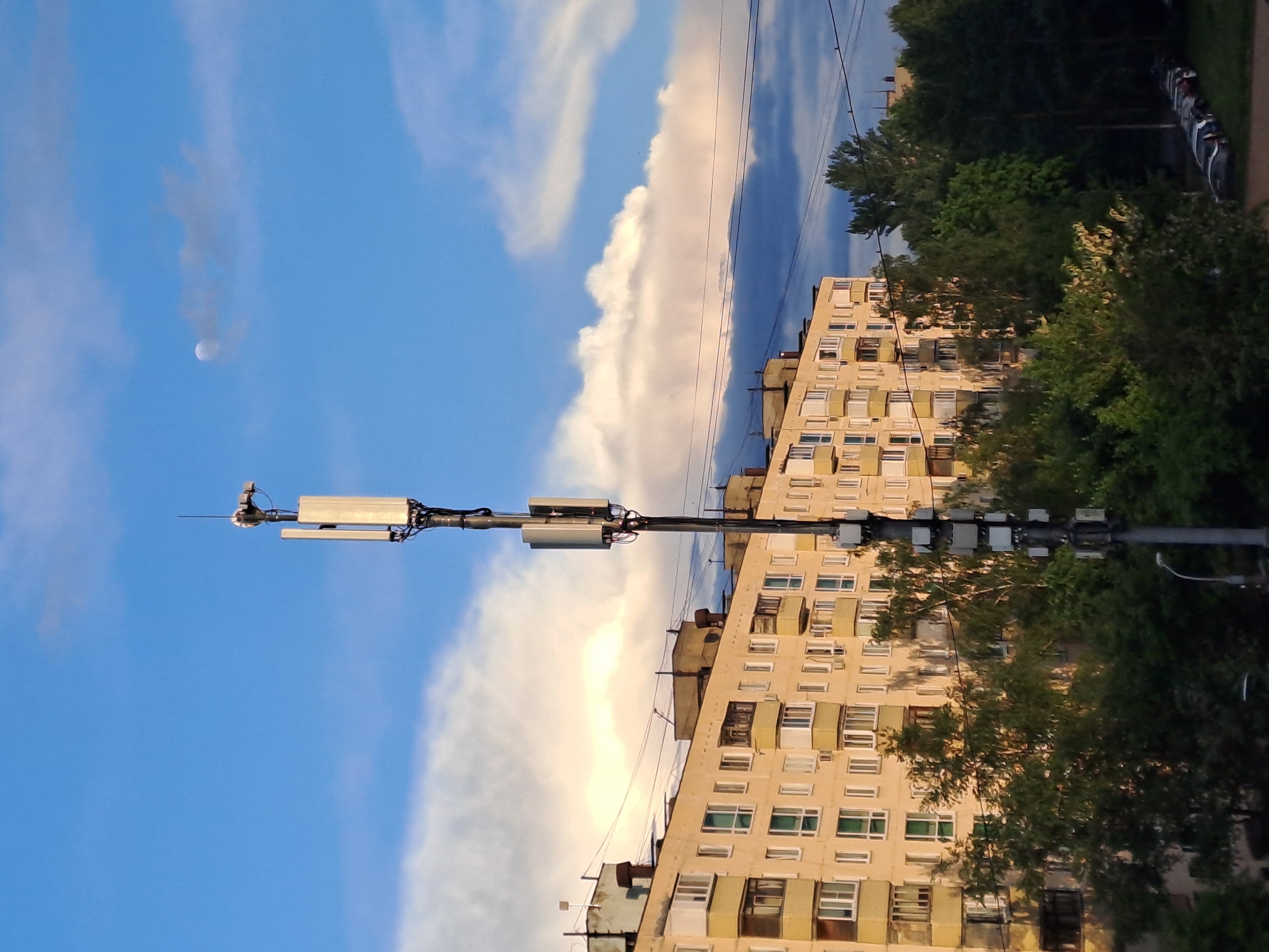 An outdoor shot in Elektrostal, Russia, shows a multi-story apartment building with rows of windows in the background, partially obscured by trees. In the foreground, there is a utility pole with antennas. A blue sky with wispy clouds and a small moon is visible. Power lines stretch across the scene. Some cars are parked near the apartment building. The image is rotated 90 degrees counterclockwise.