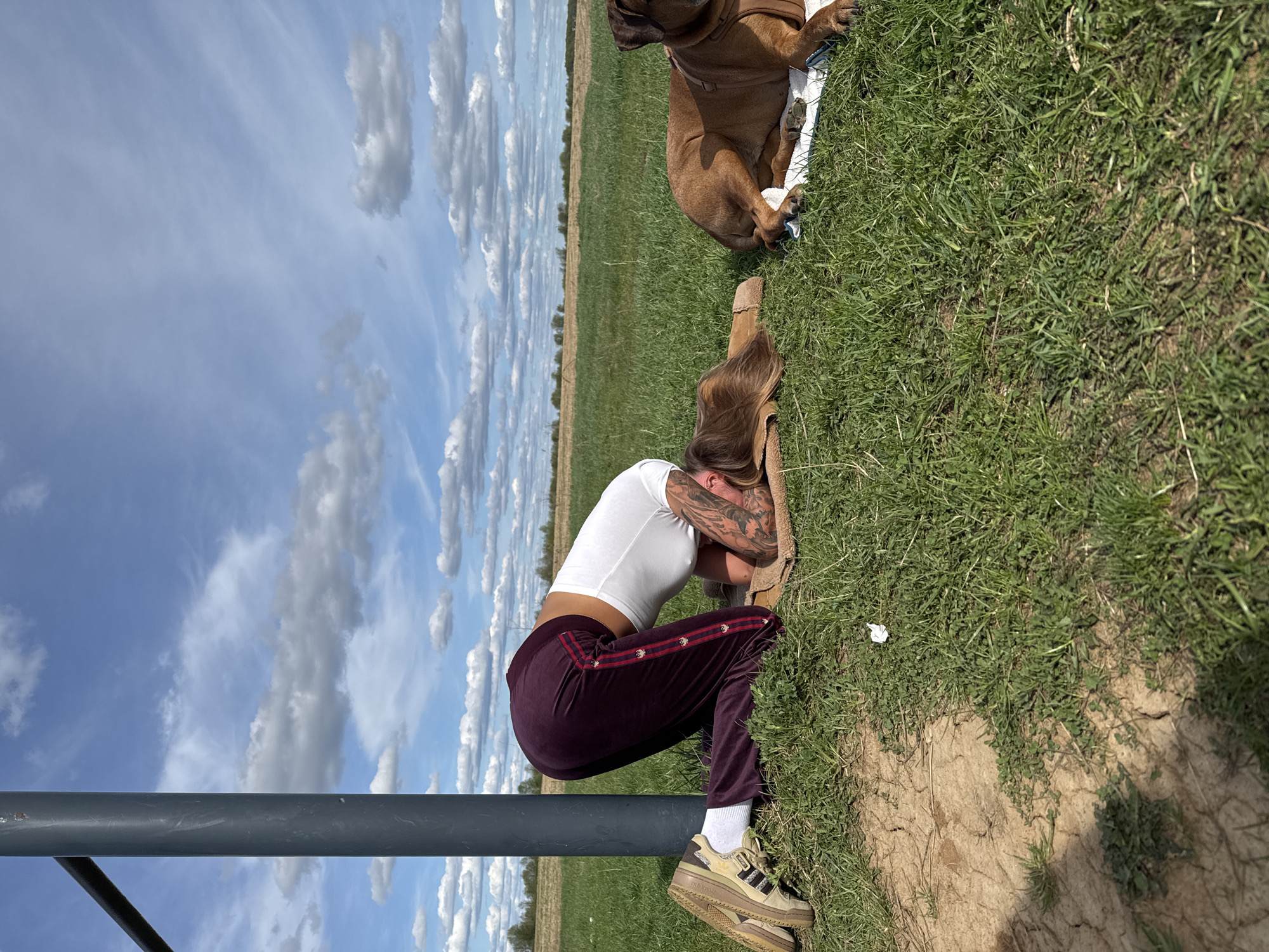 A woman wearing a white shirt, maroon track pants, white socks, and platform sneakers is squatting and resting her head on her arms near a gray metal pole. She has visible tattoos on her arm. A brown dog is lying on a blanket in the background on the grass. The setting appears to be an outdoor grassy area with a field and sky with scattered clouds in the background. The location is Ляхово, Russia.