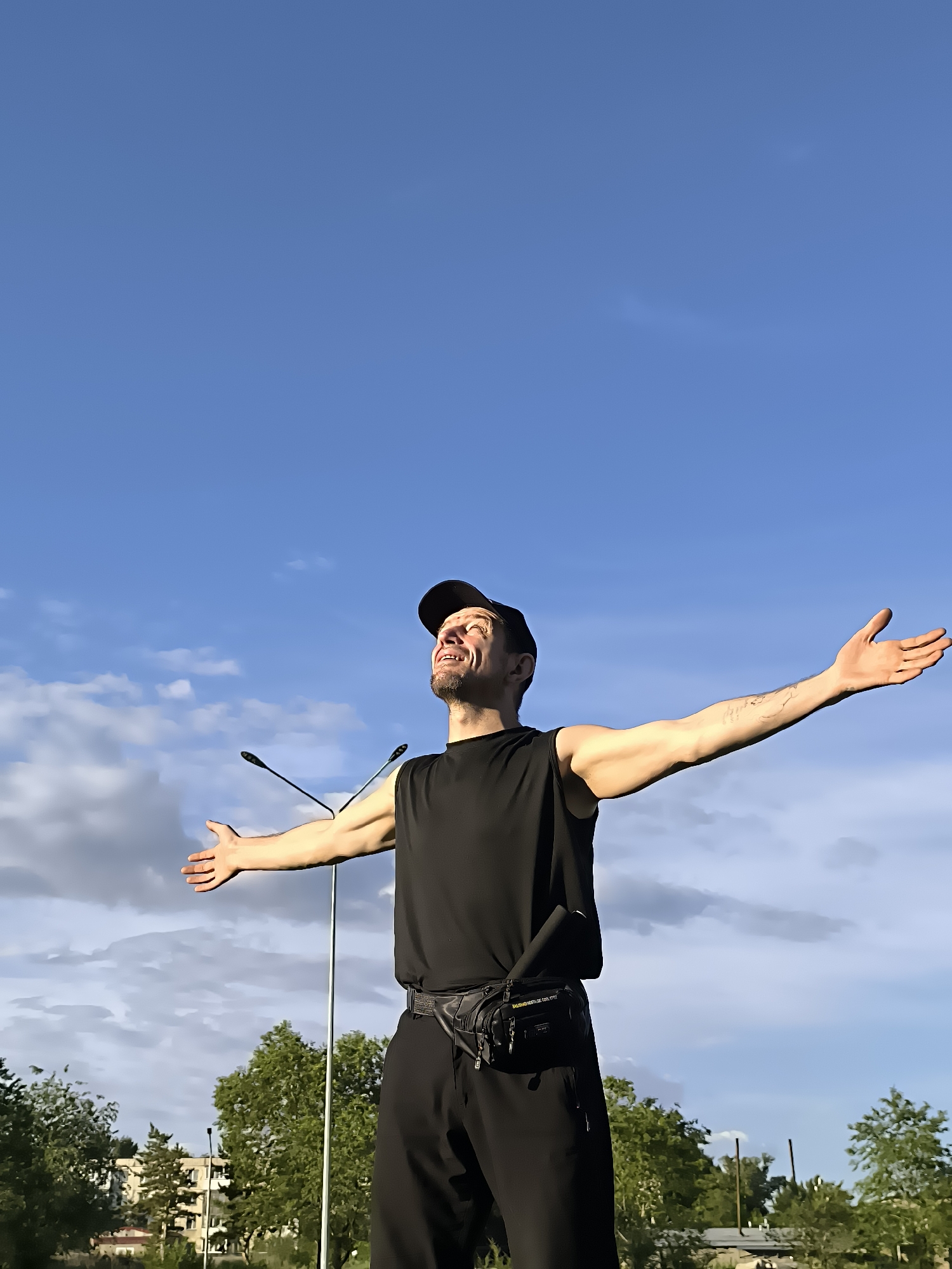 A man wearing a black sleeveless shirt, black pants, a black baseball cap, and a black fanny pack stands outside with his arms outstretched toward the sky, looking up with a smile. Behind him, the sky is blue with some scattered white clouds. Two streetlights with microphones are visible in the middle ground. Trees and buildings are in the background, suggesting an urban setting. The scene appears to be a bright, daytime environment.