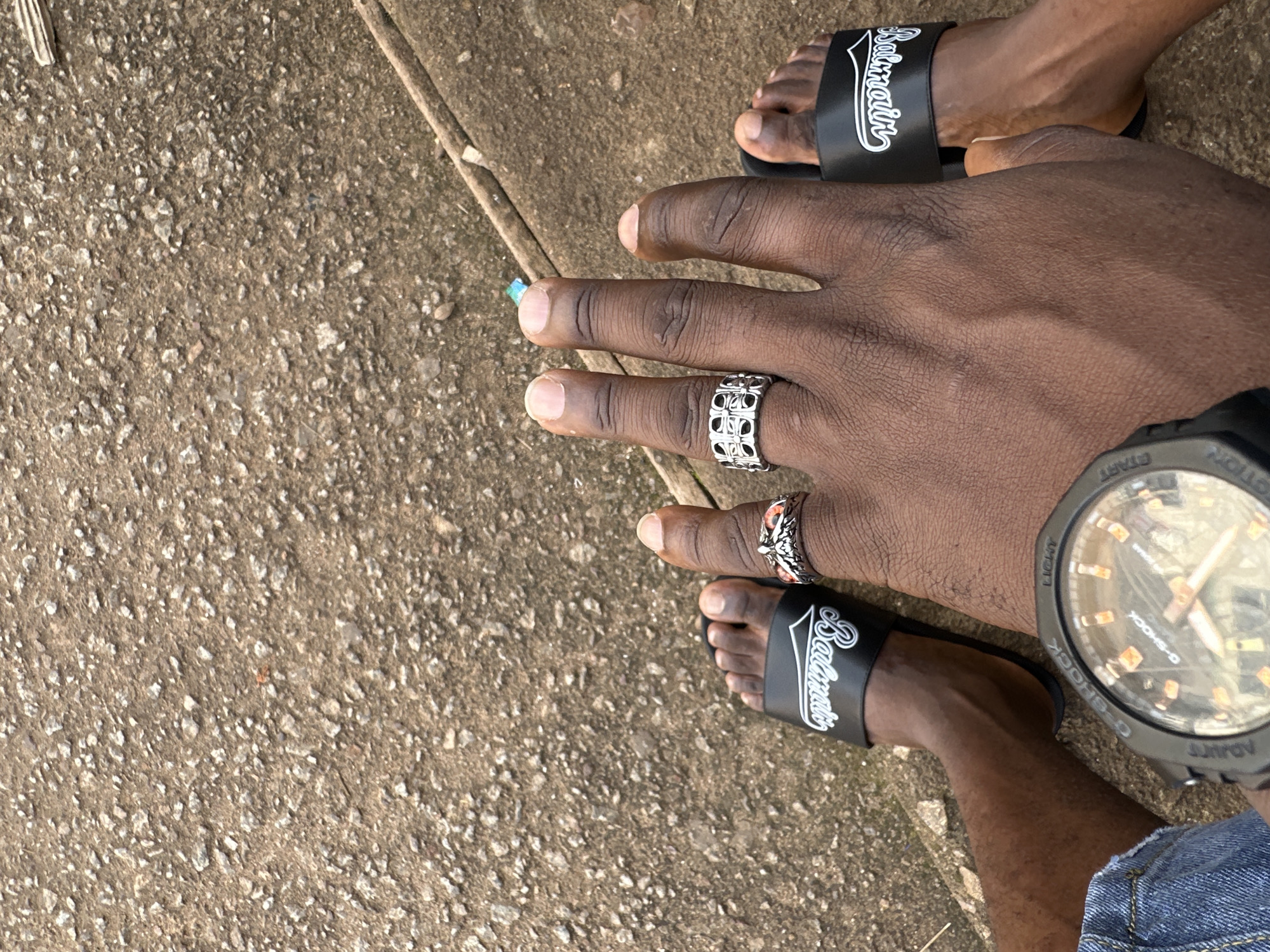 The image shows a close-up, high-angle view of a person's feet and hand resting on a gritty, brown, asphalt-like surface. The person is wearing black slides with white lettering on their feet. Their left foot has a silver ring with a red stone on the big toe. Their right hand, with several silver rings, is prominently displayed. One ring on the middle finger features a repeating pattern of squares. Another on the ring finger has a more elaborate, possibly gothic, design with a central dark stone. A large, dark digital watch with an orange-glowing face is visible on the person's left wrist. The background is a textured, pebbly surface, indicative of an outdoor urban environment. The lighting suggests daylight. There is a thin wooden stick or branch running diagonally across the upper right portion of the frame. The scene appears to be located in Bukuru, Nigeria, as indicated by the prompt.