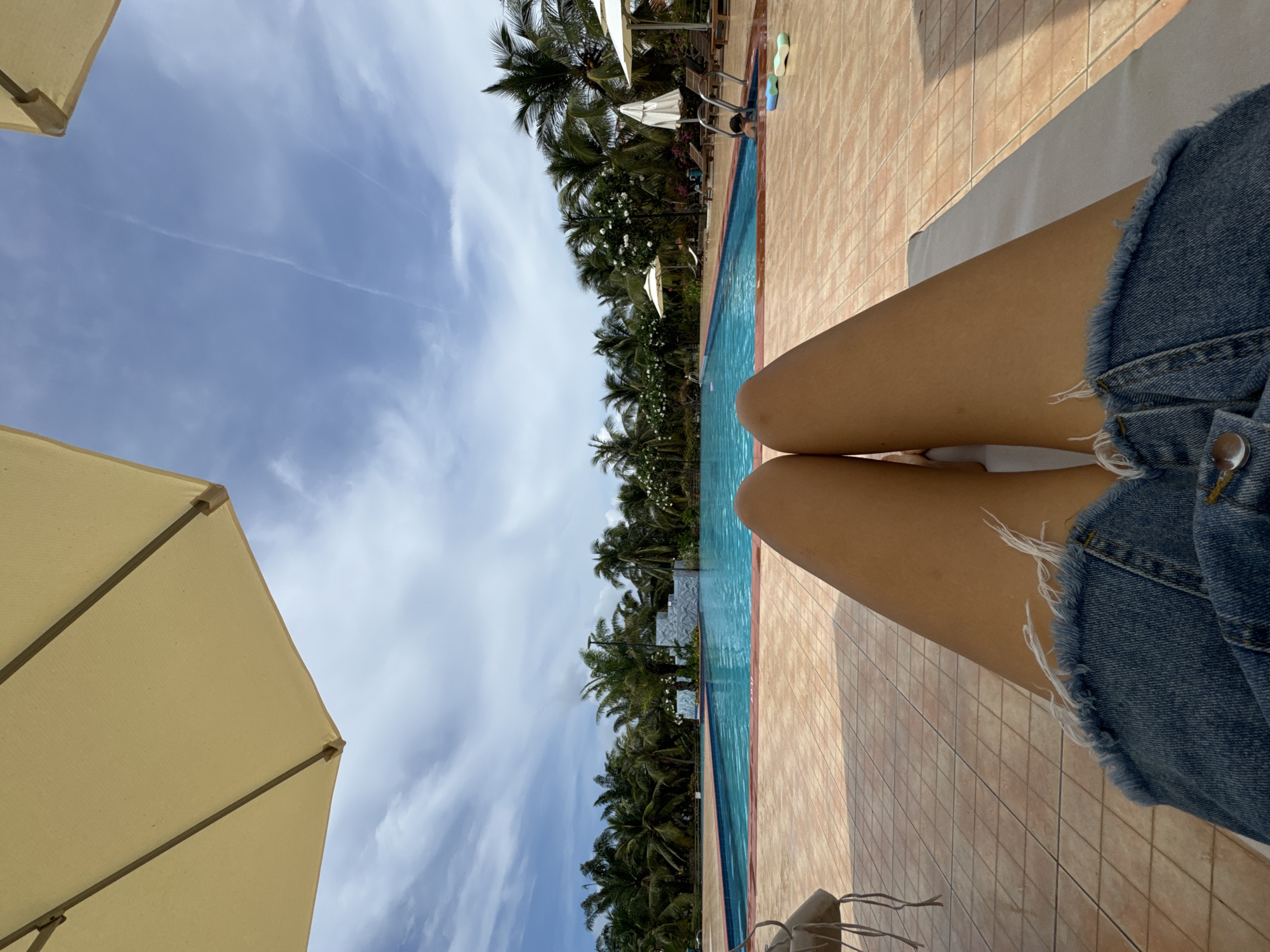 A first-person perspective shot of a person in Phu Thuy Ward, Vietnam, is sitting next to a swimming pool. The person is wearing denim shorts. An umbrella partially obscures the frame on the lower left and upper left corners. Palm trees line the other side of the pool, stretching toward a cloudy blue sky. The pool is tiled with light brown square tiles.