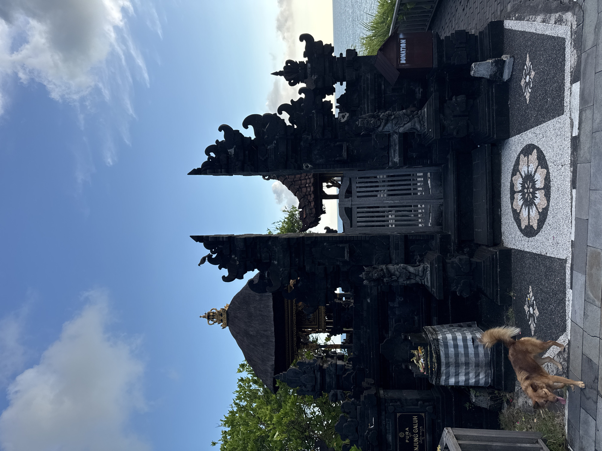 The image depicts a black stone temple structure in Kediri, Indonesia, under a blue sky with wispy clouds. The structure is ornamented with carvings, and a small donation box is visible near its upper right corner. Black and white patterned stone tiles are beneath the building, with a large floral design. A dog with light brown fur is standing in front of the temple. The ocean is visible in the background. The orientation of the image is rotated 90 degrees clockwise.