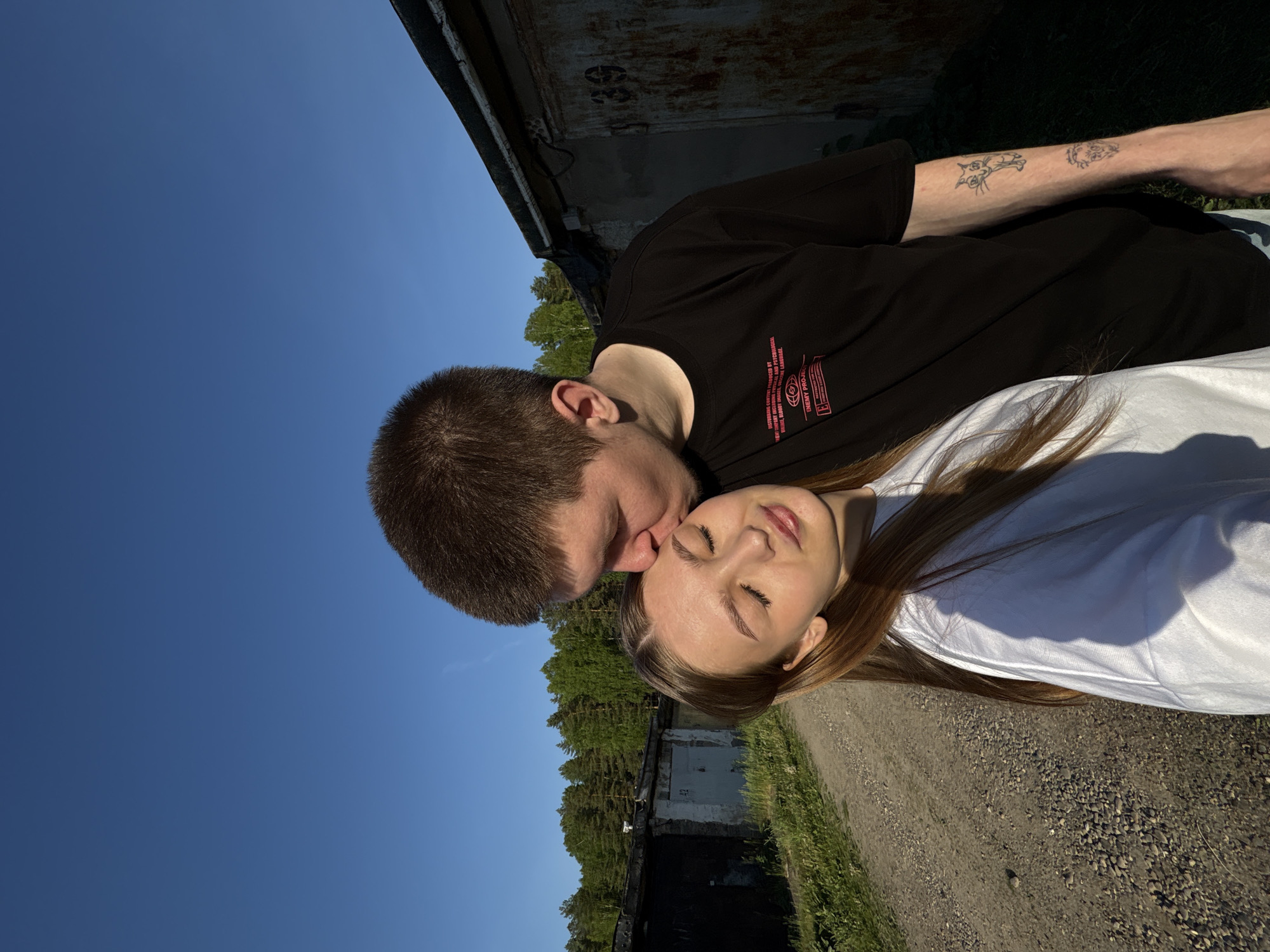 A young man with short brown hair is kissing a young woman with long blonde hair on the forehead. The man is wearing a black t-shirt with red lettering, and has tattoos on his left forearm. The woman is wearing a white t-shirt. They are standing outdoors near an overgrown gravel road and a concrete building or garage. The sky is blue. This photo was taken in the city of Chaikovsky, Russia.