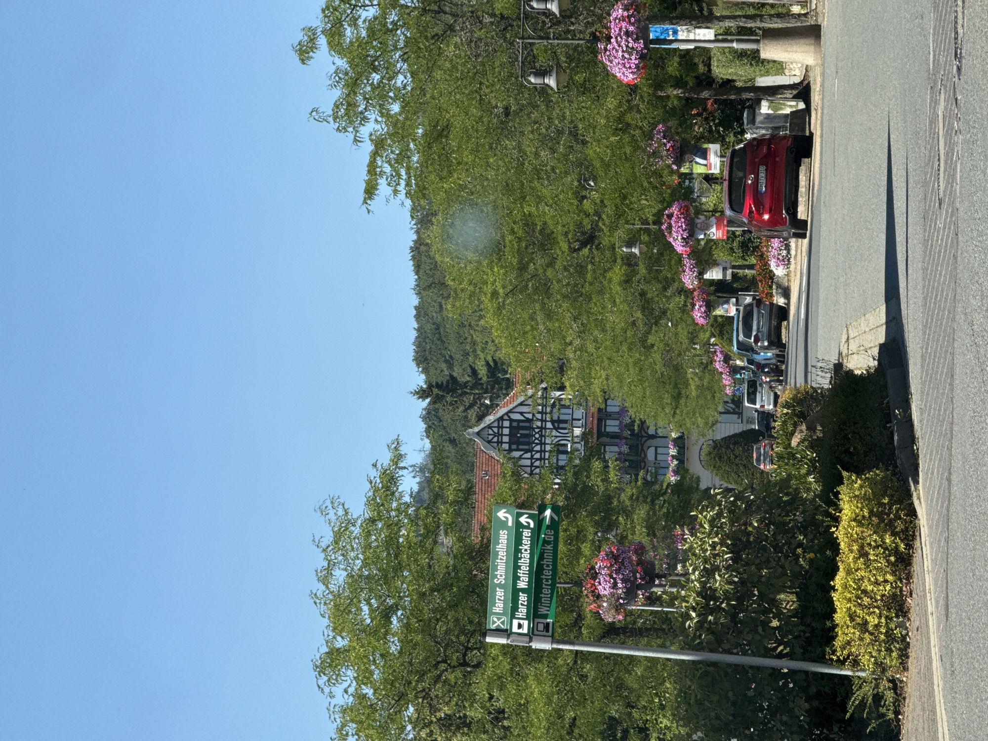 The image depicts a street scene in Bad Sachsa, Germany. A multi-story building with traditional half-timbered architecture is visible in the background. The street is lined with mature trees and decorative hanging baskets filled with pink flowers. There are several cars parked and driving on the road, including a red SUV in the foreground and a white van further down the street. A street signpost indicates directions to "Harzer Schnitzelhaus," "Harzer Waffelbäckerei," and "Wintertechnik.de." The sky is clear and blue, suggesting a sunny day.