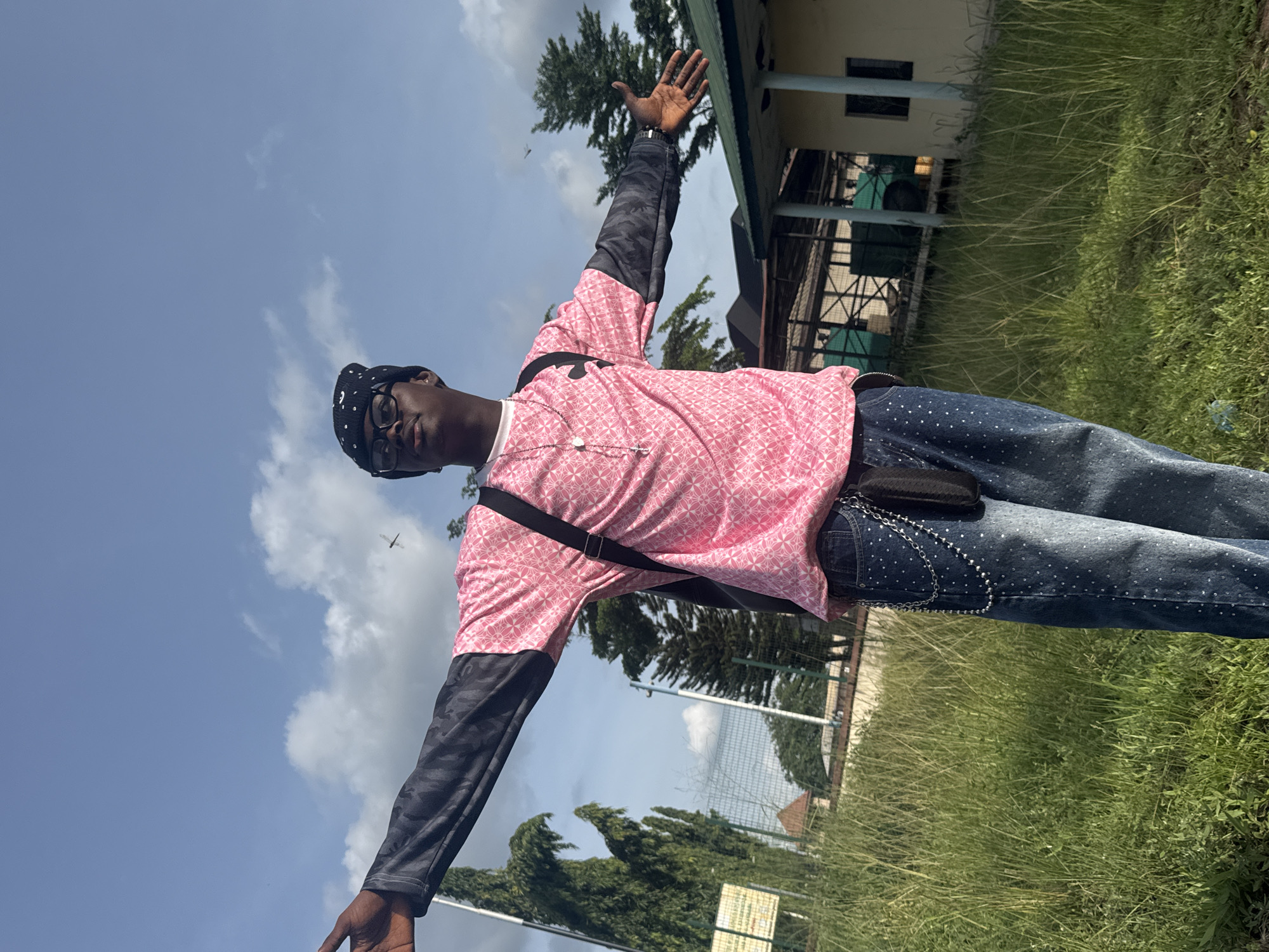 A young man stands with arms outstretched in a grassy area, possibly in Ihiagwa, Nigeria, on a sunny day. He wears a pink shirt with geometric patterns, dark-colored sleeves, dark jeans with light-colored spots, and glasses, and a black beanie. A black strap crosses his chest. He stands in front of a building, and the sky is visible in the background with scattered clouds. There are also trees in the vicinity.