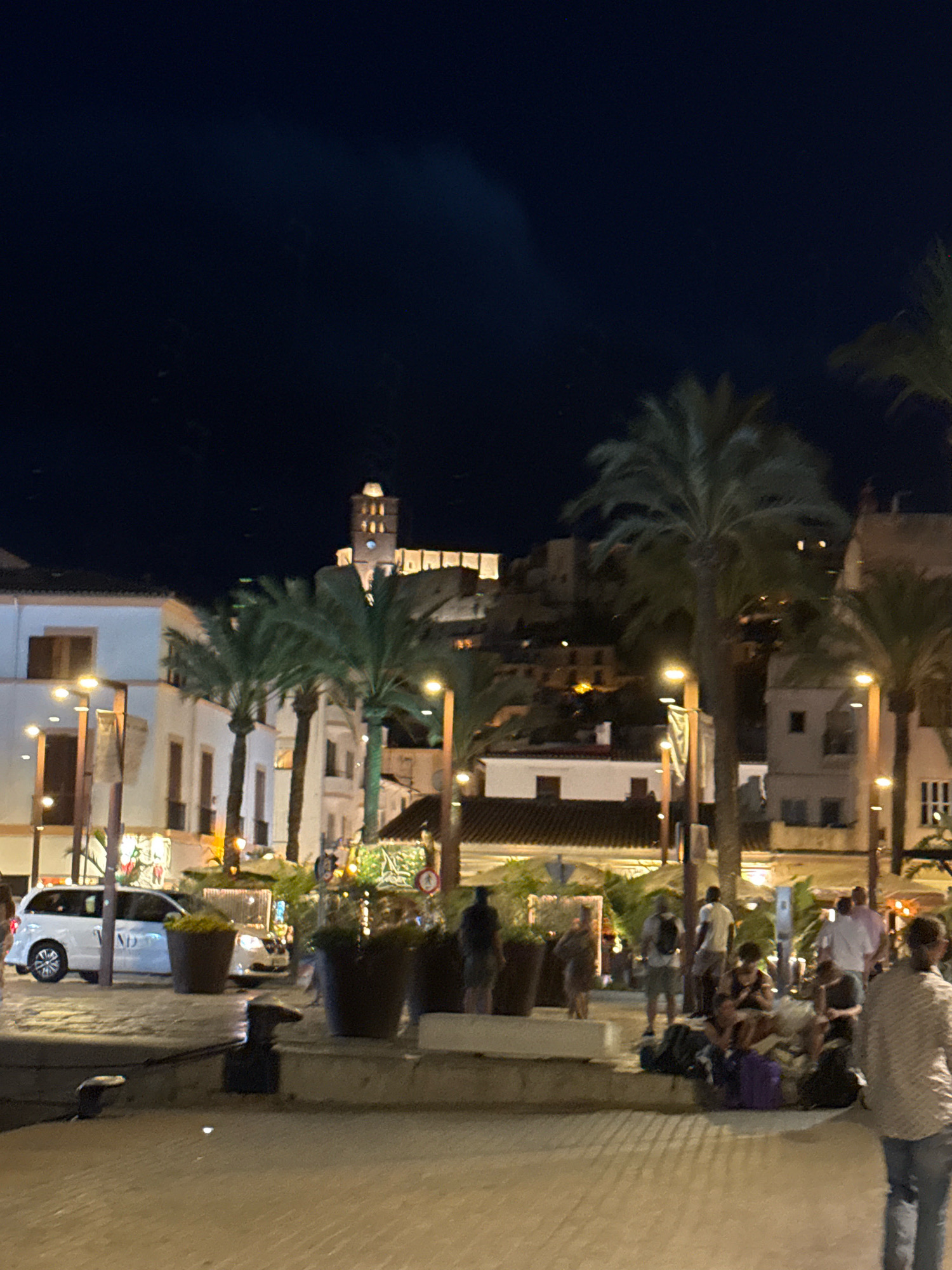 The image depicts a nighttime scene in Ibiza, Spain, likely along the waterfront or a promenade. In the background, a historic fortress or castle complex with a prominent tower is illuminated on a hillside. The foreground features palm trees lining a paved area. Several people are visible, some standing, others sitting or gathered in small groups. Luggage, including backpacks and suitcases, is scattered around the seated individuals, suggesting they may be waiting or resting. A white car is parked on the left side of the image, and in the distance, building facades with illuminated windows suggest a commercial or residential area. Streetlights are also visible, casting a warm glow on the scene. The overall atmosphere is one of a lively evening gathering in a Mediterranean setting.