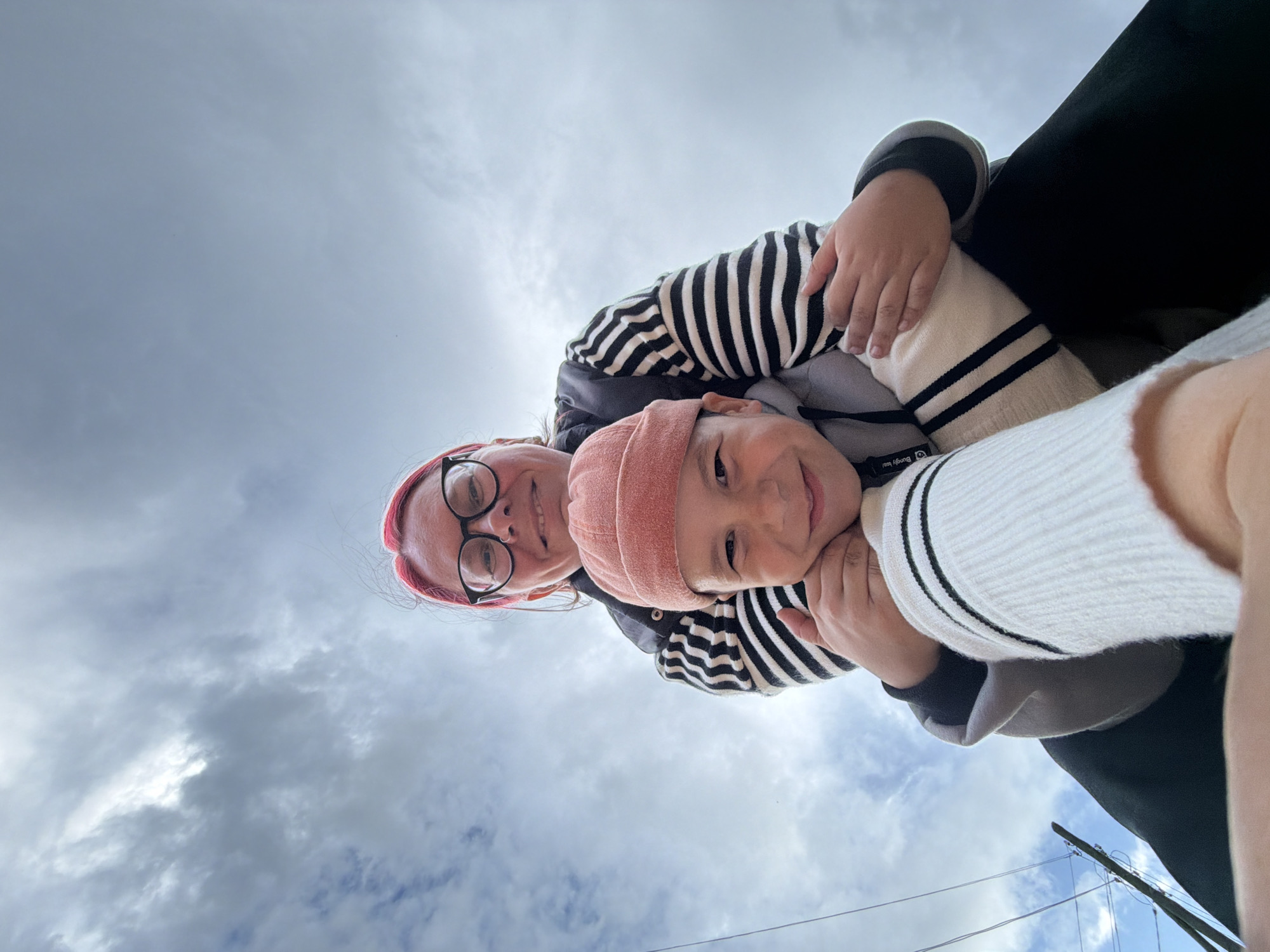 In an outdoor, low-angle shot against a cloudy sky, a person with bright pink hair and glasses is embracing a child. The person in the back is wearing a black and white striped sweater. The child, in the foreground, is wearing an orange beanie and also appears to be wearing a striped sweater. They are both looking at the camera and smiling. The backdrop includes a utility pole with wires, suggesting an urban or semi-urban environment. This scene is located in the city of Chaikovsky, Russia.