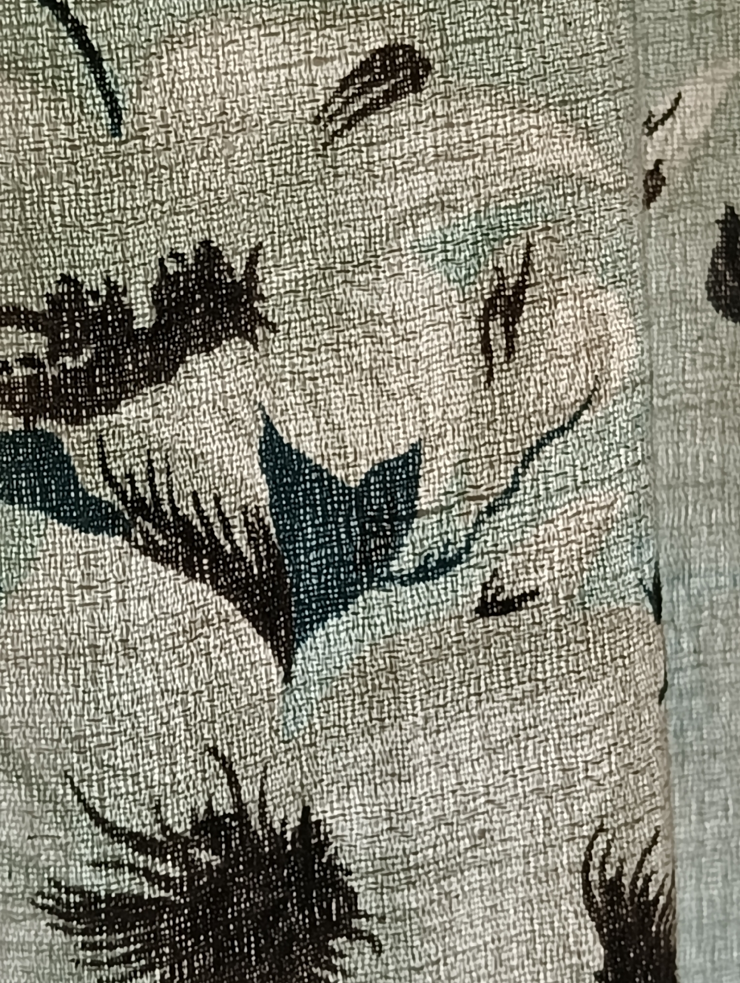 The image displays a close-up view of a patterned textile, possibly fabric for upholstery, curtains, or clothing. The primary subject is the fabric itself, which features a distinct woven texture, resembling tweed or a similar material, in a light grey or beige base color. Scattered across this background are stylized patterns in dark blue, black, and hints of a lighter blue-green. These patterns appear abstract but suggest natural forms, such as leaves, branches, or perhaps fragmented floral motifs, with some elements having a spiky or feathery appearance.
There are no people or discernible activities depicted. The scene is a static, detailed view of the fabric's surface. On the right side of the main image and in some cropped sections, a vertical fold or seam is visible, indicating the material is draped or shaped, possibly forming a part of an object like a curtain. The lighting is even, and there are no specific cues for time of day or weather. No text is visible on the fabric. The overall aesthetic is one of muted colors and organic, non-representational designs on a textured surface. The location context provided is Biliaivka, Ukraine.
