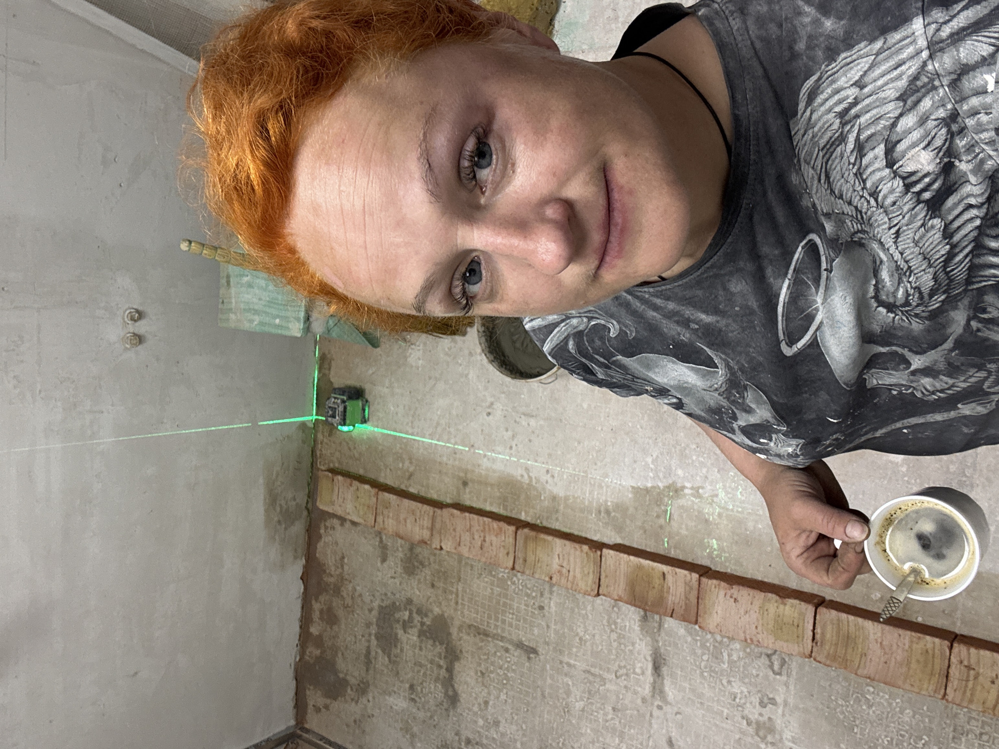 The image depicts a woman with red hair, fair skin, and blue eyes in an interior space under construction, likely in the city of Chaikovsky, Russia. She is wearing a grey t-shirt with a skull graphic and holding a white mug of coffee. A green laser level is emitting a horizontal beam across a row of bricks that are set against a wall of unfinished grey plaster. The room appears to be in the process of renovation.