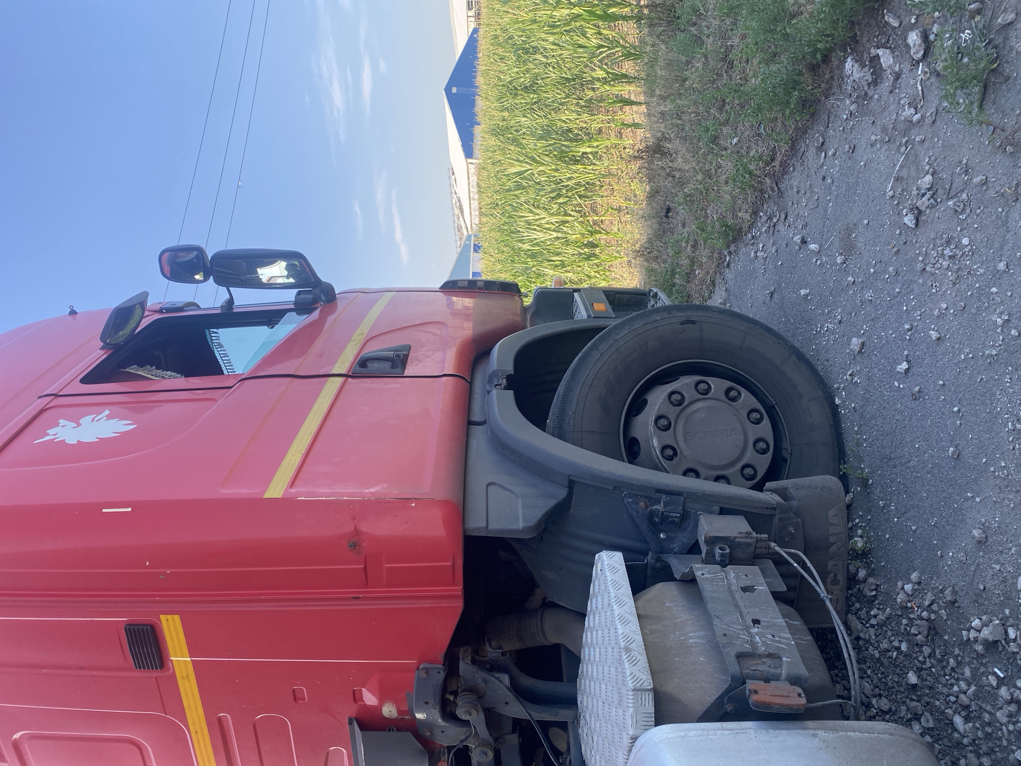 A red Scania truck is parked on the side of a road next to a cornfield. The truck is partially visible, with its front wheel and side of the cab showing. The cab is red with a yellow stripe and a silver lion emblem on the side. The truck is parked on an asphalt road with gravel on the side. In the background, there is a cornfield with green stalks of corn. Beyond the cornfield, there are two large blue and white buildings with white roofs. The sky is clear and blue with a few wispy clouds. The location is in Mais'ke, Ukraine.