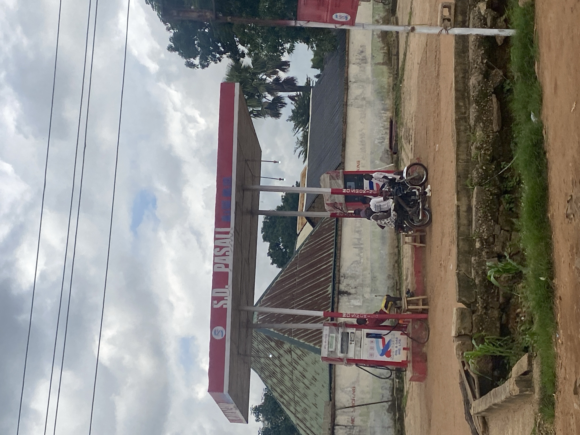 The image depicts an outdoor scene at a gas station in Tataura, Nigeria. A red and white gas station canopy, labeled "S.D. PASALI", is the central focus. Beneath the canopy, a red gas pump stands with visible labels, and a person in a red shirt and yellow shorts is seated on a stool beside it. In front of the gas pump, a motorcycle with three individuals is parked. Two are seated on the motorcycle, and one is standing. To the right of the motorcycle, another person is standing, and a second stool is visible. Behind the gas station, structures with corrugated metal roofs are present. The ground is unpaved dirt, and a stone and concrete drainage ditch with some vegetation is in the foreground. Overhead, power lines are strung across a cloudy sky. The overall impression is of a functional, somewhat rustic, local service station in a rural or semi-urban Nigerian setting.