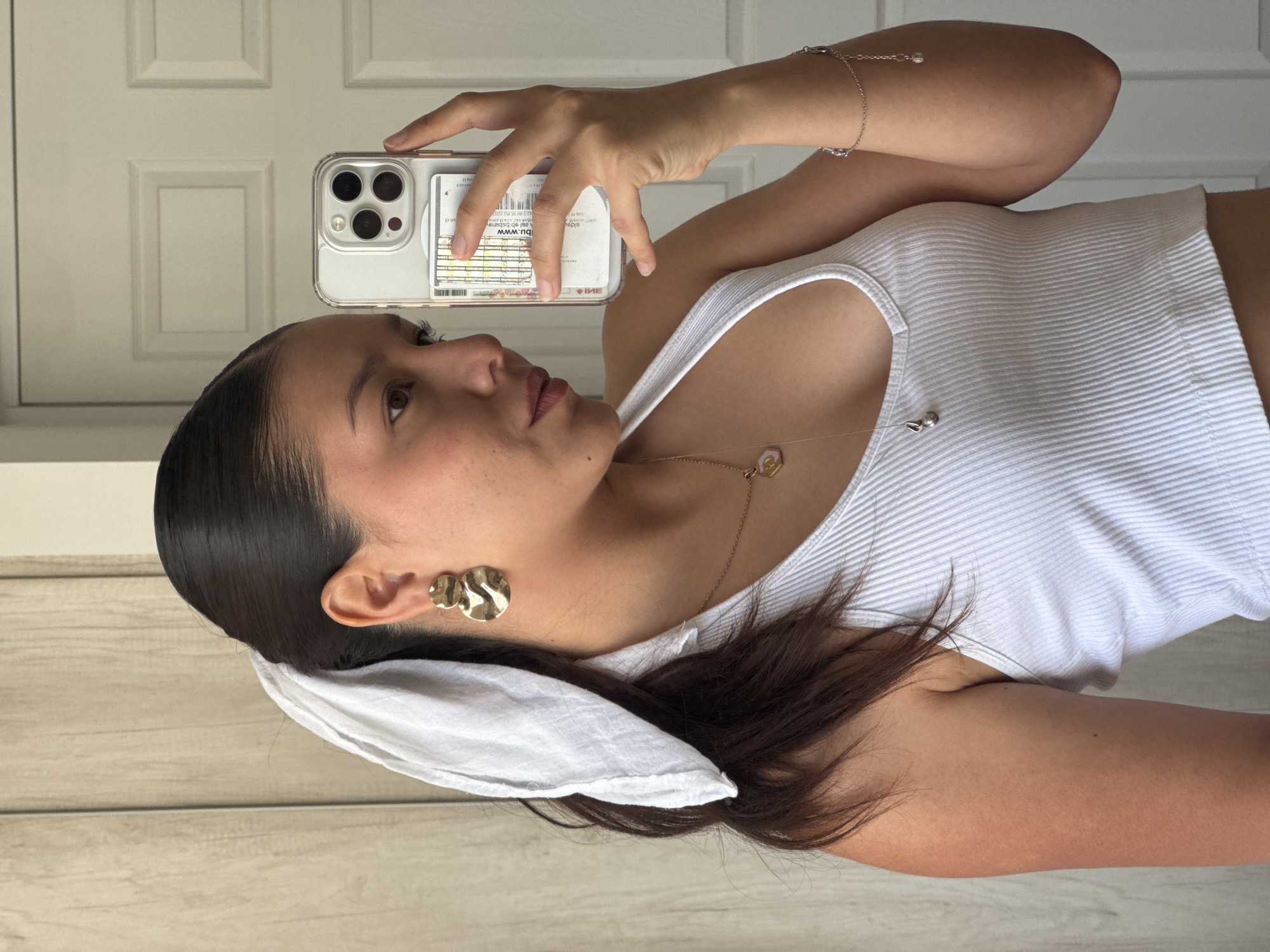 A woman is holding a phone up to take a picture of herself. She is wearing a white ribbed tank top and a gold necklace. Her hair is pulled back with a white scarf. She has gold earrings on. The phone is in a clear case with a card tucked in the back. The background appears to be a white door.