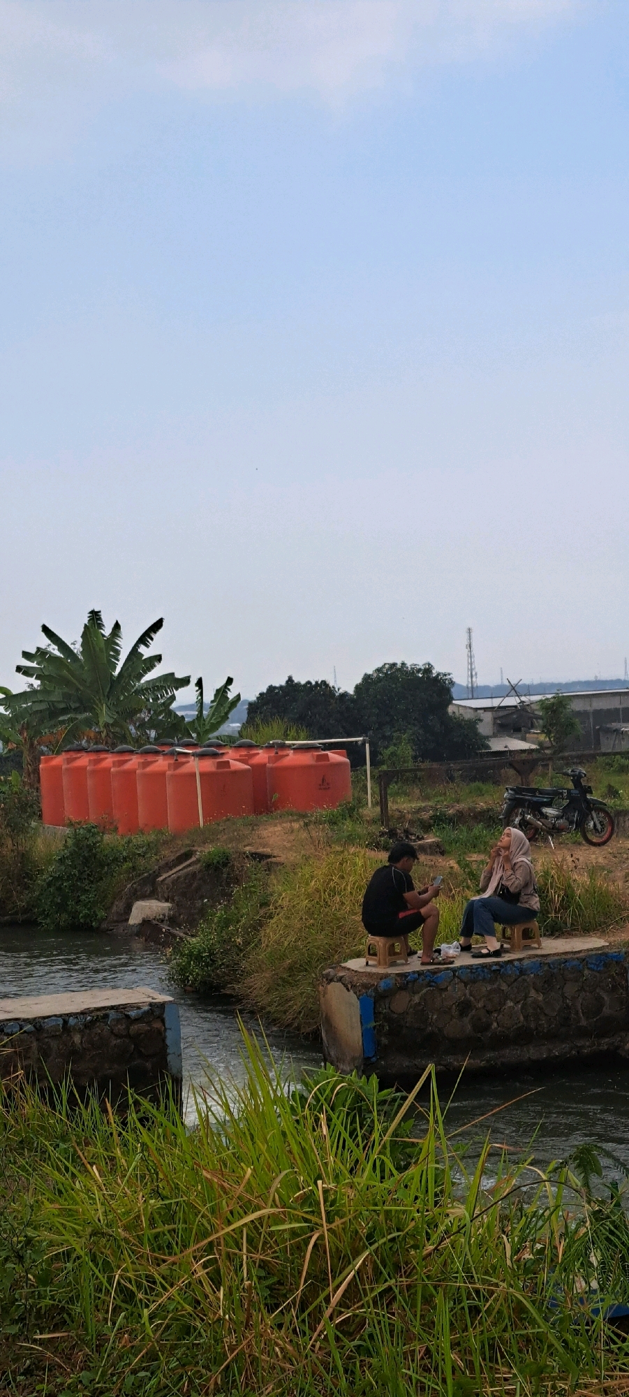 In Sumber Gedang, Indonesia, an outdoor scene depicts a tranquil setting with a focus on natural elements and human interaction. The foreground is dominated by lush green grass and a flowing stream, with a stone and concrete structure bordering the water. Beyond the stream, a raised platform made of stone and concrete serves as a resting spot for two individuals.
A man, seated on a small plastic stool, wears a black t-shirt and dark shorts. He is looking at his phone. Beside him, a woman, wearing a hijab and a light-colored jacket over blue jeans, also sits on a small plastic stool. She is looking upwards, and her hands are near her face. Between them, on a plate, are what appear to be snacks.
In the middle ground, a row of large, orange water tanks is visible, suggesting agricultural or industrial use. Behind the tanks, banana trees with large leaves stand prominently, adding to the tropical atmosphere. The background reveals a mix of trees and what appears to be industrial structures, including a tall tower and power lines, indicating the proximity of developed areas. A parked motorcycle is also visible to the right of the water tanks. The sky is a clear, pale blue with faint white clouds, suggesting a daytime scene, likely with moderate weather conditions.