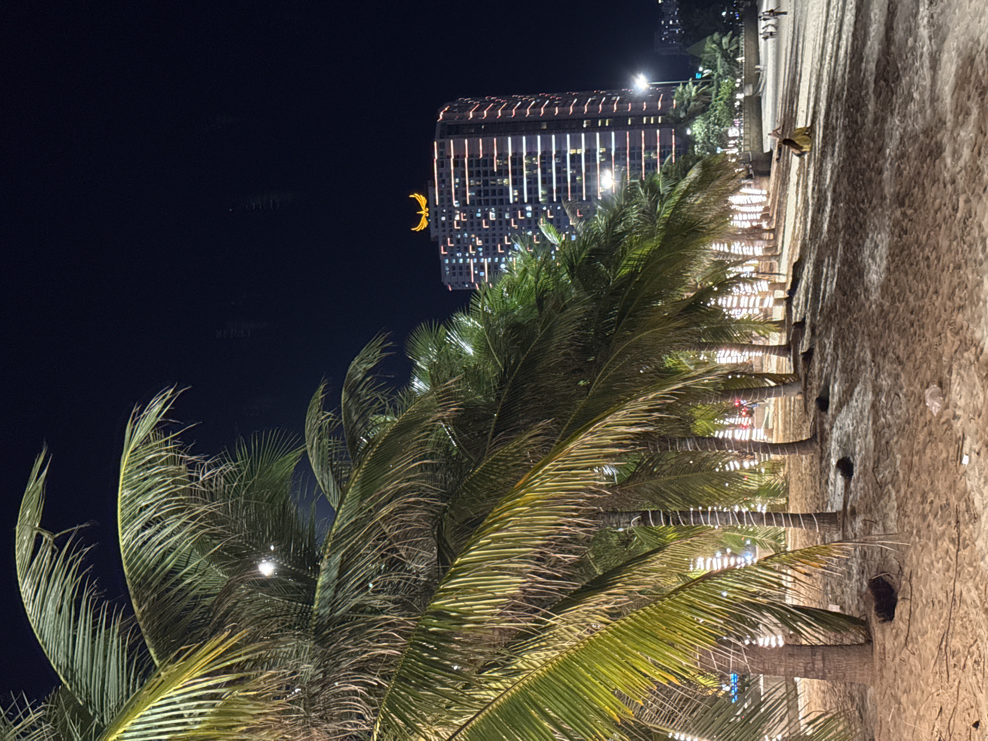 This is a nighttime, full-frame, eye-level shot of a sandy beach in Nha Trang, Vietnam, likely taken from a resort area. In the foreground, a row of palm trees with their fronds illuminated by decorative lights line the beach. The sand is a light brown color, with some areas appearing wet. In the mid-ground, several people are visible walking and sitting on the beach. Further in the background, a tall, modern building is brightly lit with vertical stripes of white and pink lights. A golden, winged logo is prominent at the top of this building. The sky is dark and clear, indicating a cloudless night. The overall atmosphere is serene and festive, suggesting a popular tourist destination.