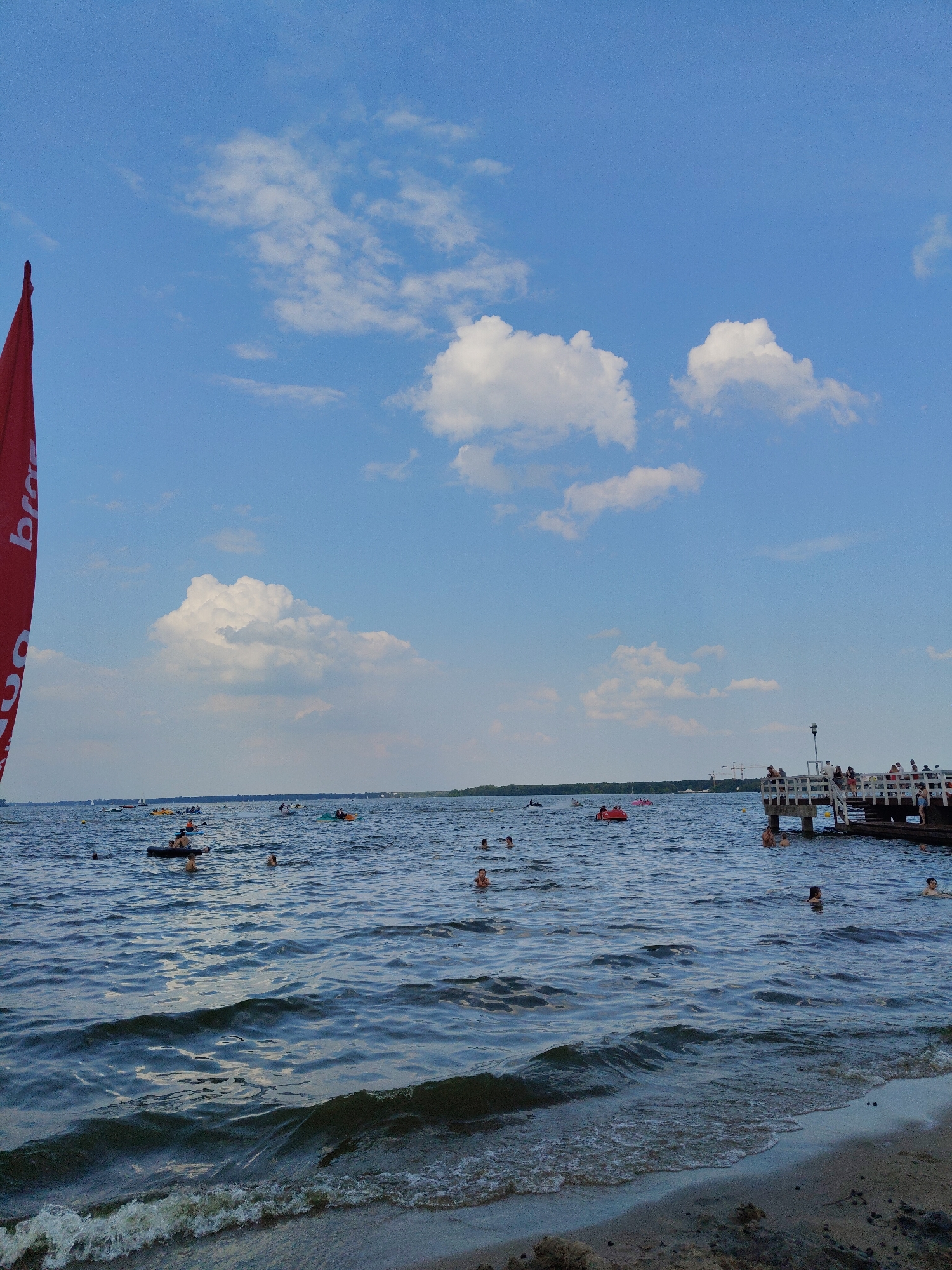 The image depicts a sunny day at a beach and lake in Nieporęt, Poland. A bright red flag with white lettering is visible on the left side of the frame, partially obscuring the blue sky filled with scattered white clouds. The water is populated with numerous individuals swimming, floating on inflatable devices, and engaging in various water activities. A wooden pier extends into the lake on the right side of the image, with several people standing on it, observing the water or socializing. The shoreline in the foreground is sandy, with small waves lapping at the edge. In the background, a tree line and some distant structures suggest the presence of a town or resort area. The overall atmosphere is one of leisure and recreation on a warm summer day.