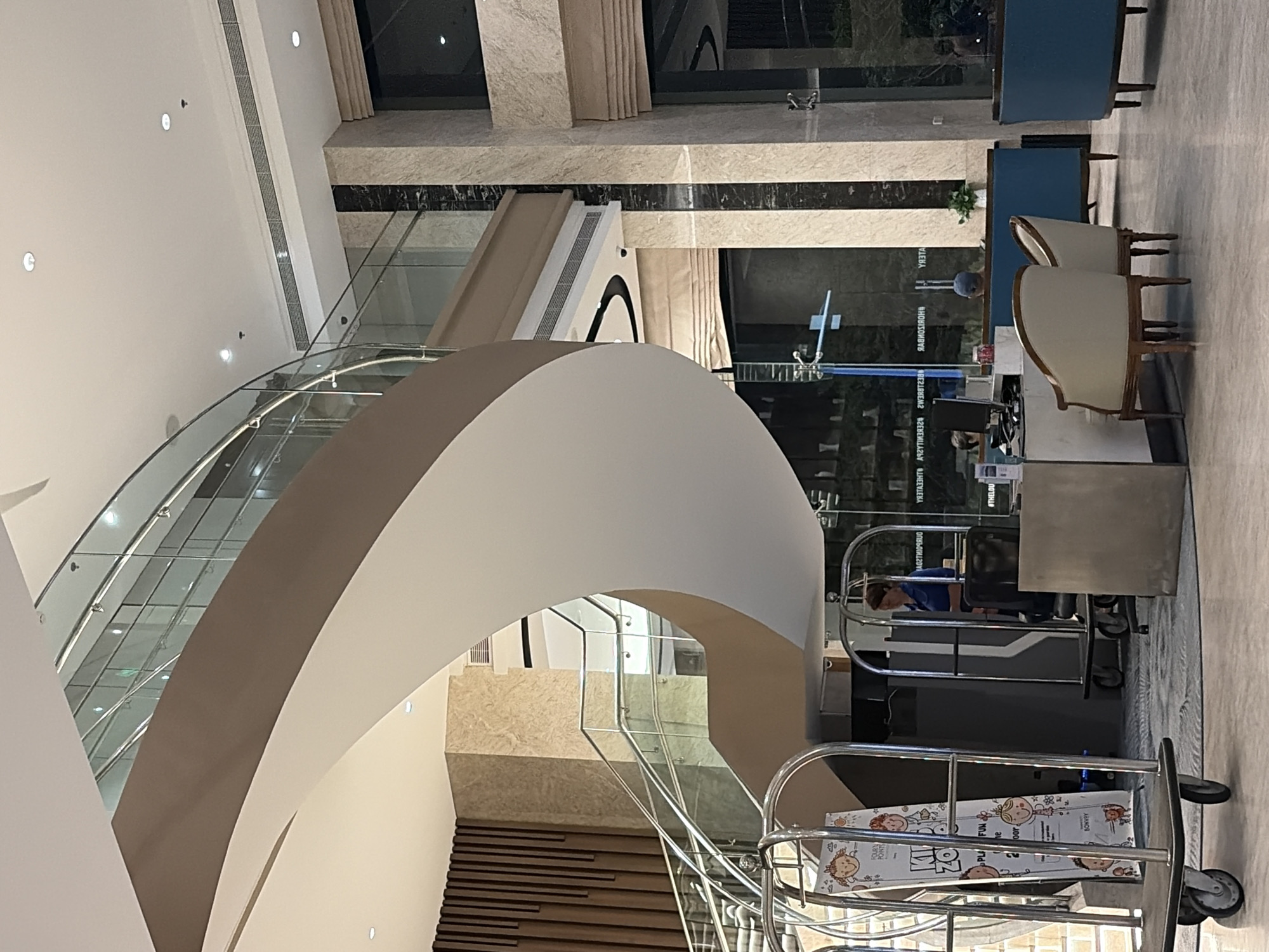 The image captures the interior of a modern hotel lobby in Da Nang, Vietnam. A prominent feature is a grand, sweeping spiral staircase with a white and brown railing, leading up to the second floor. The lobby has a polished marble floor and is decorated with warm, earthy tones and stone accents. In the foreground, a luggage cart displays a colorful poster for a "Kids Zone" activity. To the right, a reception desk is manned by a person in a blue uniform, with a computer and seating area visible. Further back, lounge chairs and blue seating furniture suggest a comfortable waiting or gathering space. Natural light filters in through large windows, illuminating the elegant design of the lobby.