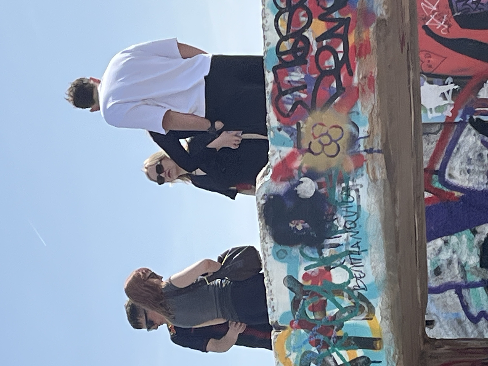 A bright, sunlit outdoor scene in Barcelona, Spain, features several individuals interacting with a large, graffiti-covered concrete structure. The image is taken from a low angle, looking upwards towards a clear blue sky.
The main subjects are four people positioned near or on the robust, vertical concrete structure, which is heavily adorned with vibrant, multi-colored graffiti. The graffiti includes a mix of abstract designs, bold letters, and various symbols in red, blue, green, yellow, black, and white.
In the upper part of the image, a person with dark hair, wearing a white t-shirt and black trousers, stands with their back to the camera, seemingly looking downwards or across the structure. Below them, a person with blonde hair, wearing sunglasses and a black top, looks towards the left side of the frame. Further down and to the left, another person with reddish-brown hair, dressed in a dark top, is partially visible, also looking away from the camera. A fourth person's head and shoulders are partially seen below them. The individuals appear to be observing the surroundings or posing.
Notable details include the bright, sunny weather with a clear blue sky, indicative of daytime. A faint white contrail from an airplane is visible high in the sky on the left. The concrete structure itself is a prominent feature, suggesting an urban or industrial backdrop transformed by street art. Within the graffiti, some text is discernible; for instance, the word "DEMIANQUE" (or a similar spelling) is written vertically in black against a light blue and white background on the right side of the structure. The overall impression is one of casual exploration or enjoyment of the public art in an outdoor setting.
