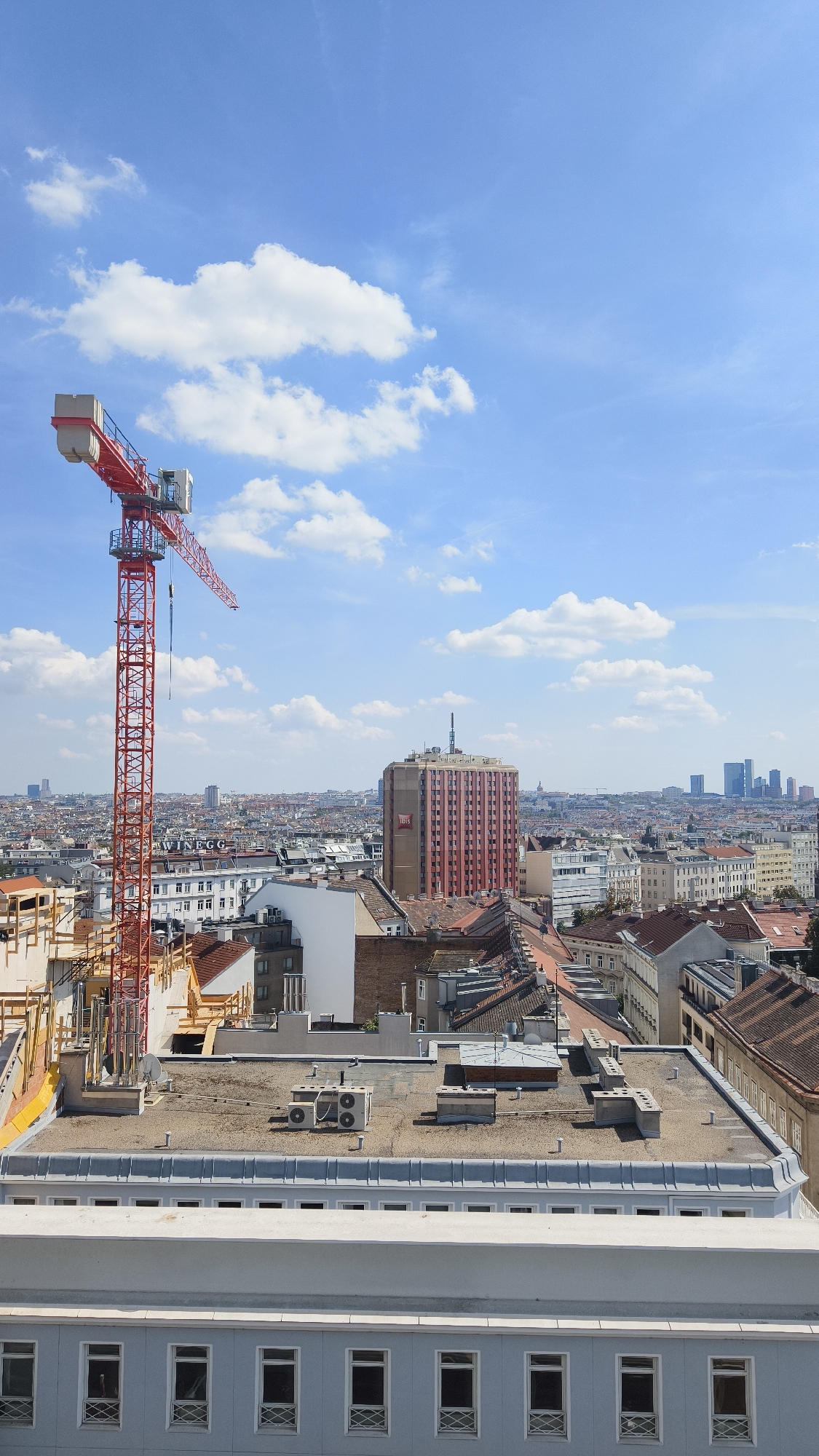 The image captures an urban skyline in Vienna, Austria, under a bright blue sky with scattered white clouds. Dominating the left side is a tall, red construction crane, its arm extended horizontally over the city. In the mid-ground, a distinctive pink, multi-story building with vertical brown stripes stands prominently. Several other buildings of varying heights and architectural styles, mostly with terracotta tiled roofs, fill the landscape. The foreground features the flat rooftops of several structures, including one with visible air conditioning units and ventilation systems. The lower portion of the image shows the facade of a light blue building with a series of evenly spaced windows. The scene appears to be a clear, sunny day, suggesting an outdoor environment at an elevated vantage point overlooking the city.