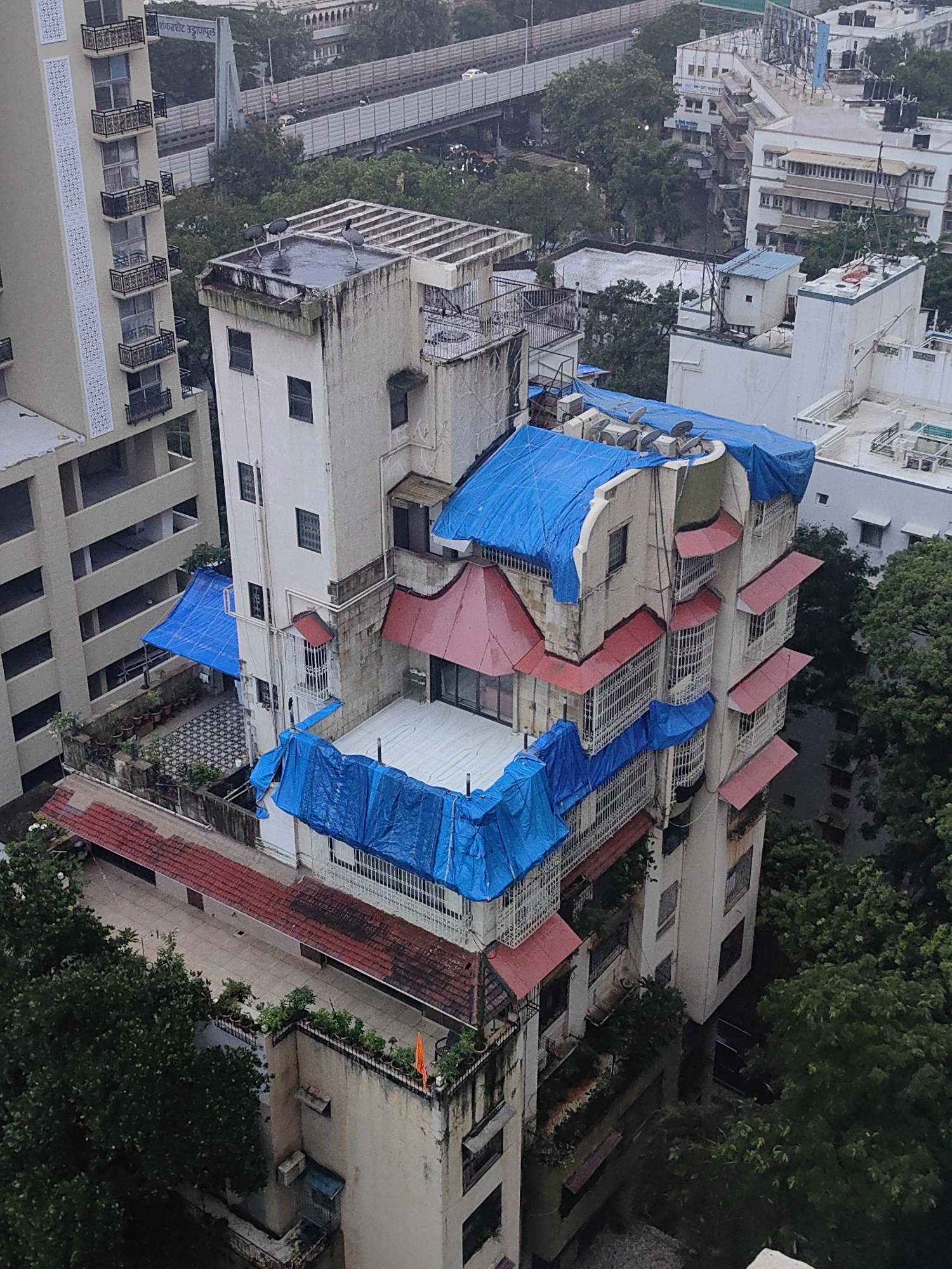 This is an aerial shot of a dense urban environment in Mumbai, India. The scene is dominated by a cluster of multi-story buildings, some appearing to be residential apartments. Several of these buildings have been covered with blue tarpaulins, likely for protection against the elements or ongoing construction. The architecture is typical of Mumbai, featuring balconies with decorative ironwork and red-tiled sloped roofs on some sections.
In the foreground, a building with several balconies is partially covered by blue tarpaulins. Adjacent to it, a taller, lighter-colored building with a distinctive patterned facade is visible. Another building, seemingly older, has multiple levels with red-tiled roofs and numerous windows with intricate metal grills. Blue tarps are also spread across different levels of this structure. A small, orange flag is visible on the rooftop of a lower section of the building.
In the background, a highway overpass can be seen with cars driving on it. The surrounding area is filled with lush green trees, indicating the presence of parks or natural vegetation within the city. The overall impression is one of a busy, built-up area of Mumbai, where construction and weather protection measures are clearly visible.